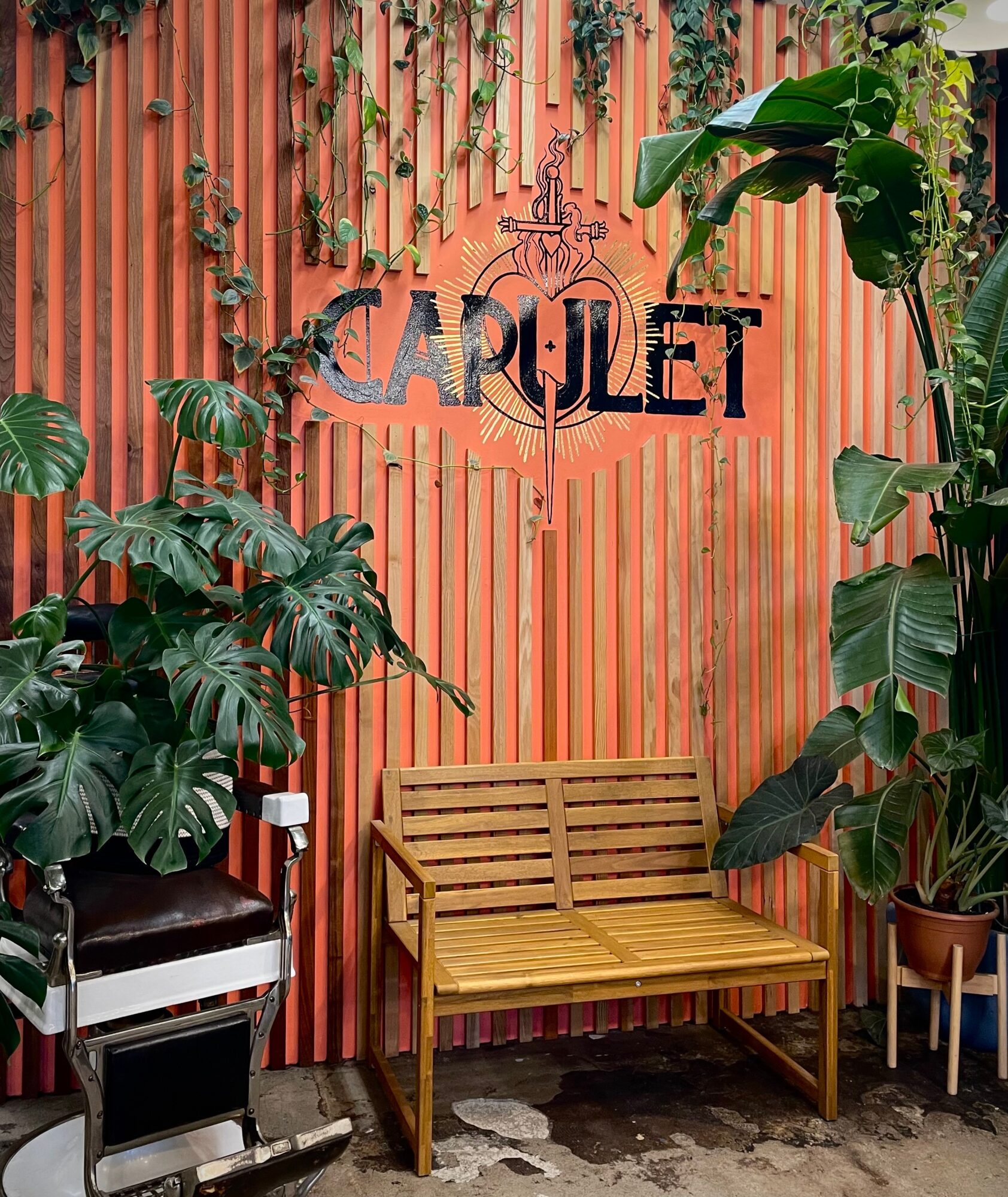 Wooden bench in front of a pink striped wall with green plants and a sign reading 'CAPULET'.