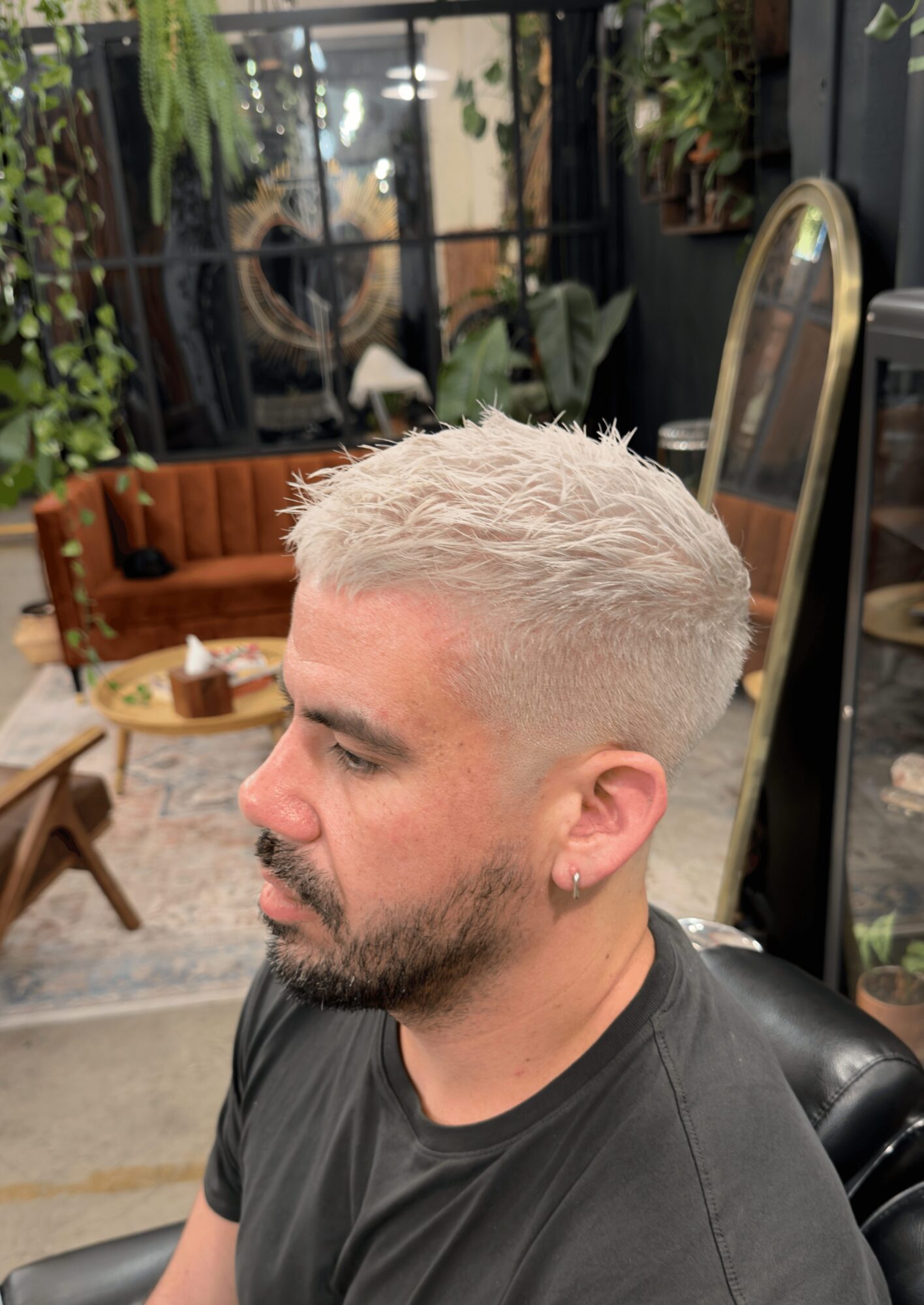 Side profile of a man with platinum blonde hair and a beard, sitting indoors with plants and furniture in the background.
