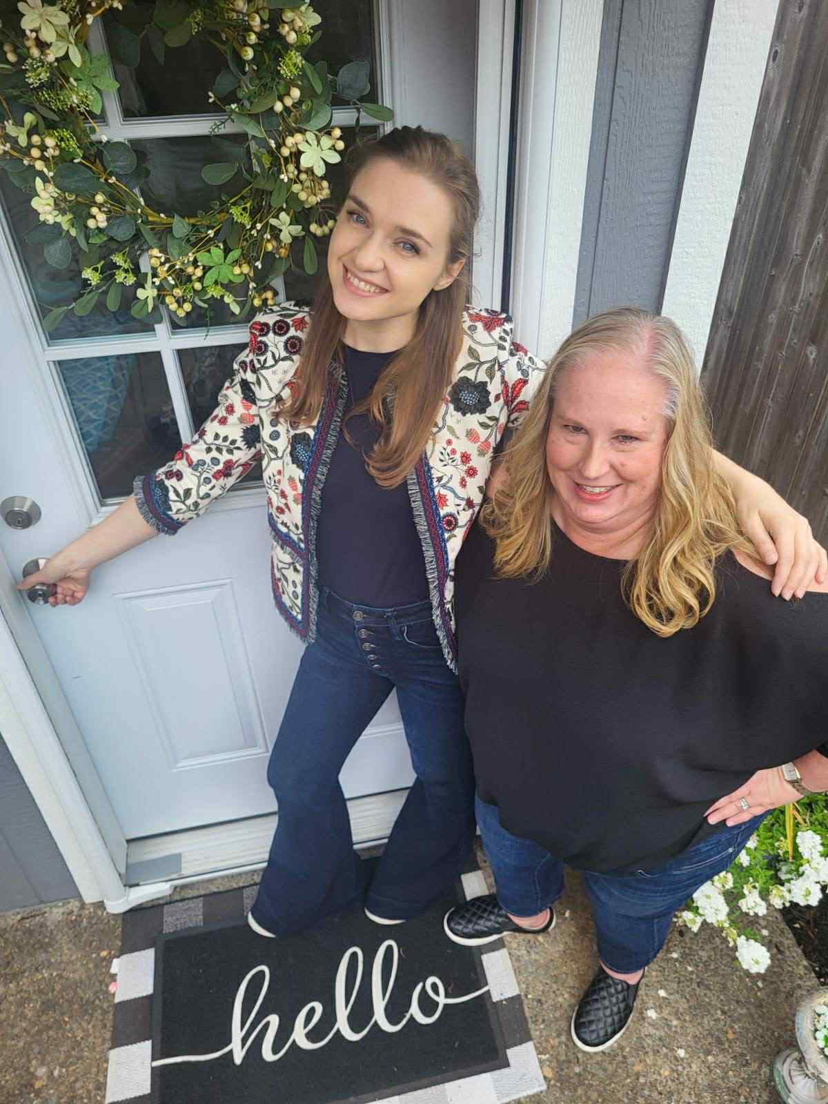 Two women smiling outside a door with a welcome mat, one with arm around the other, greenery in background.