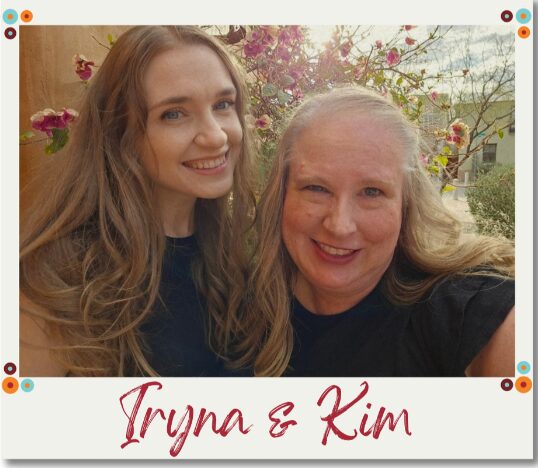 Two women smiling outdoors with blooming trees in the background, one with long hair and the other with light hair, wearing dark clothing.