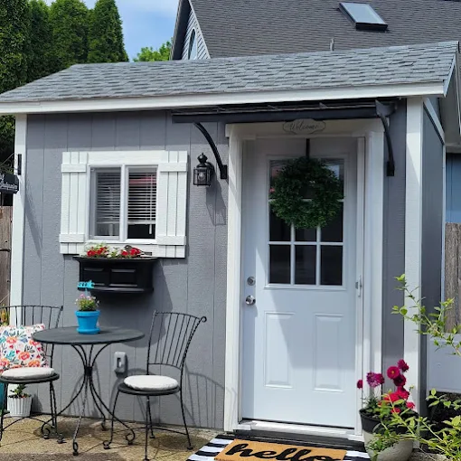 Gray house exterior with white door, window with shutters, hanging wreath, outdoor table, chairs, and potted plants.