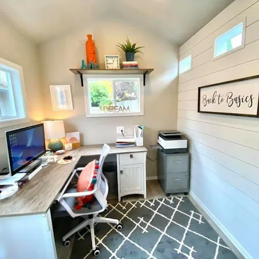 Home office with desk, chair, computer, shelves, and wall decor, illuminated by natural light from windows.