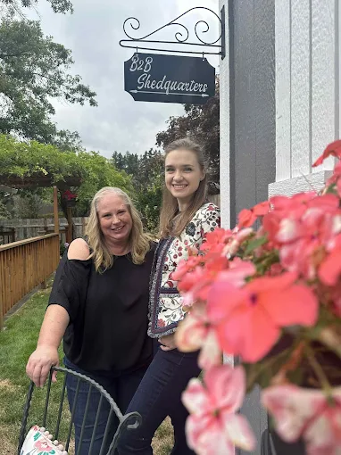 Two women smiling outdoors near a building with a sign that reads 'Shedquarters,' with pink flowers in foreground.