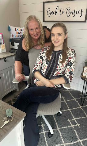Two women smiling in a room with a sign that says 'Back to Basics' on the wall, sitting on a chair.