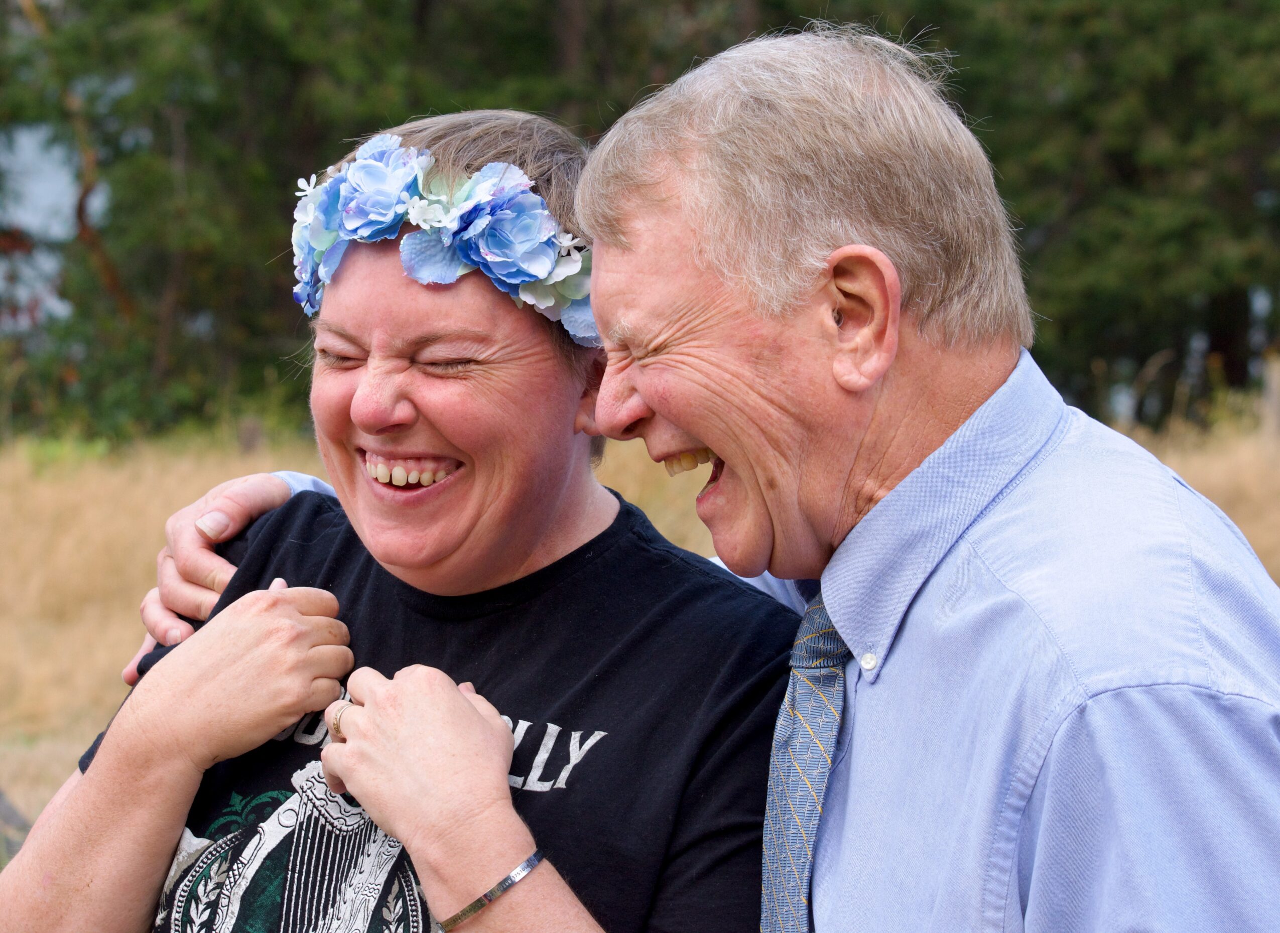 Two people laughing outdoors, one wearing a flower crown, the other in a light blue shirt, embracing each other.