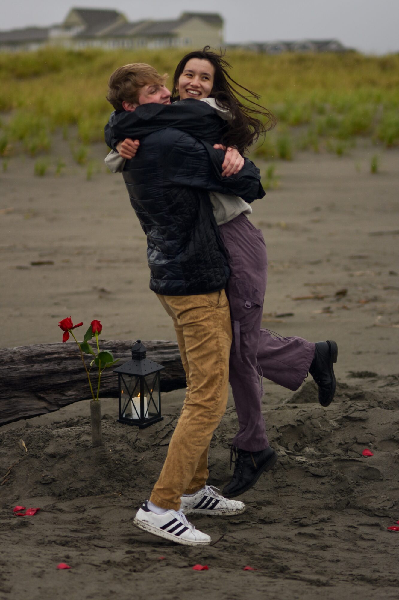 Two people hugging on a sandy beach, smiling, with a grassy area and houses in the background.