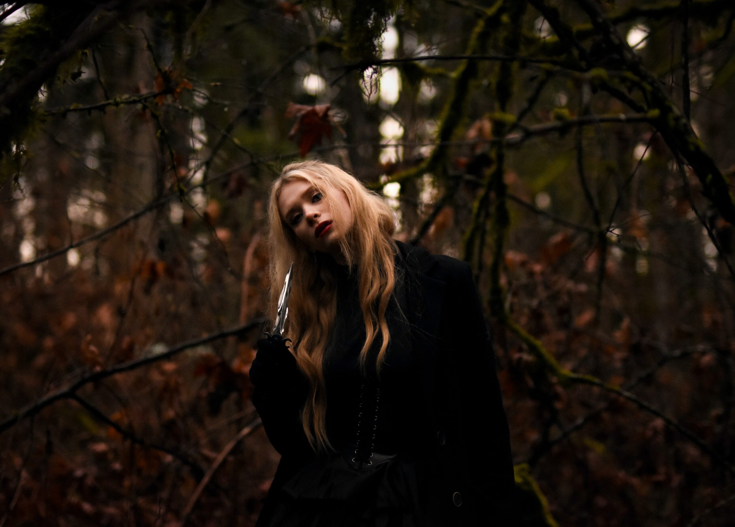 Young woman with long blonde hair stands in a dark, wooded area with bare branches and fallen leaves.