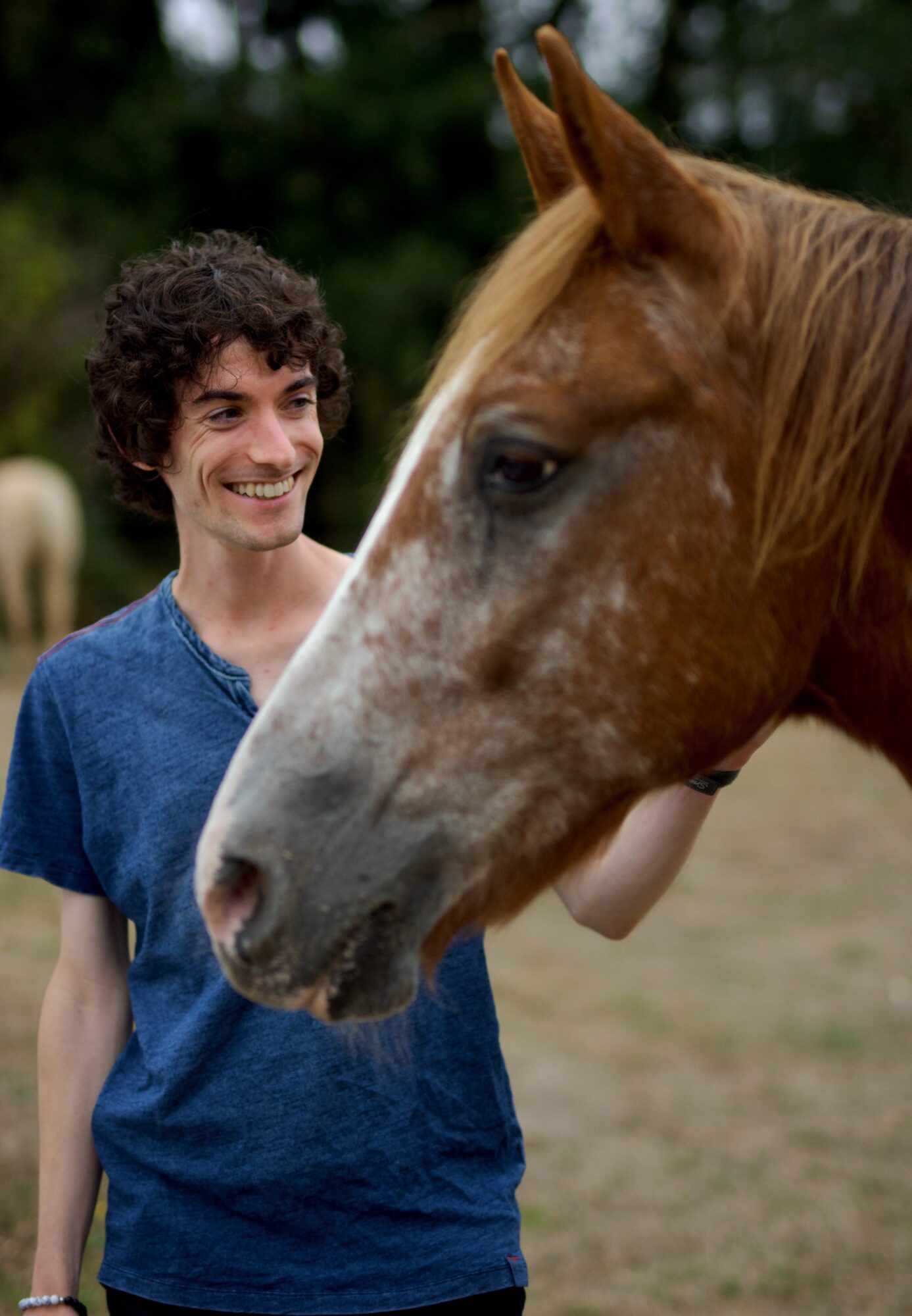 Person with curly hair smiling near a horse outdoors, background with trees and another horse, daytime.
