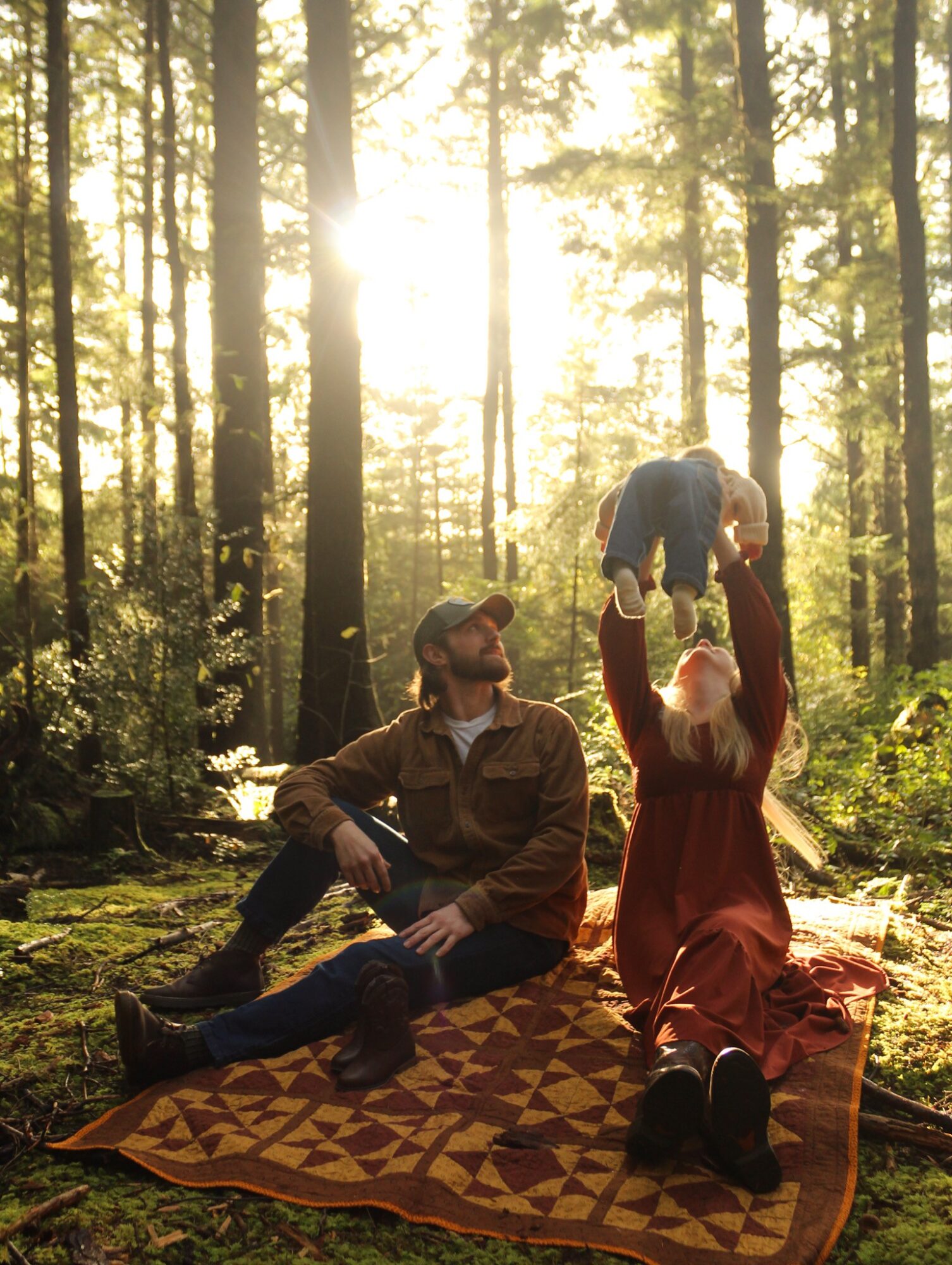 A man and a woman sit on a blanket in a forest, with sunlight filtering through trees; the woman lifts a child.