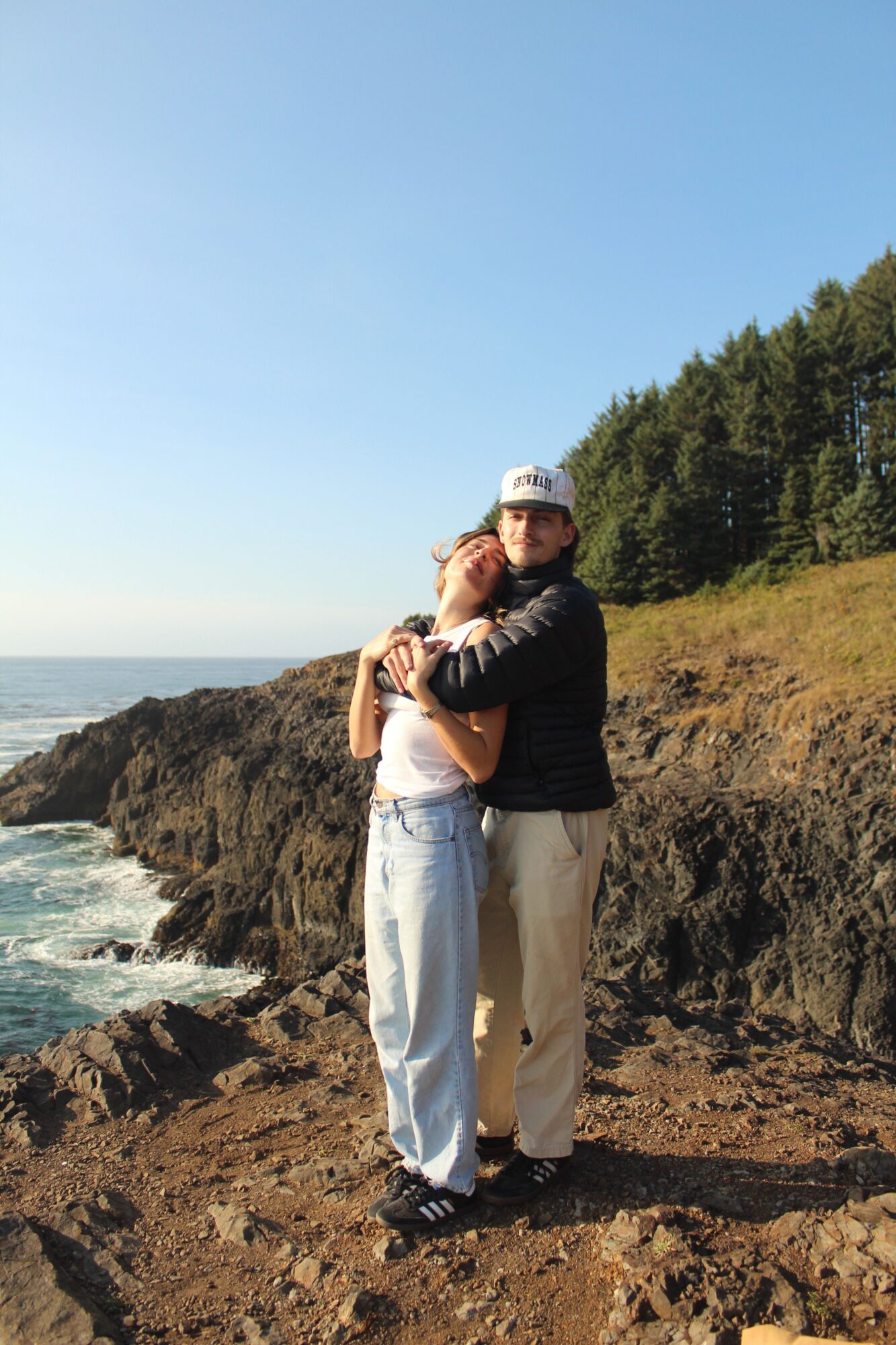 Two people hugging on a rocky coastline with trees and ocean in the background, under a clear blue sky.