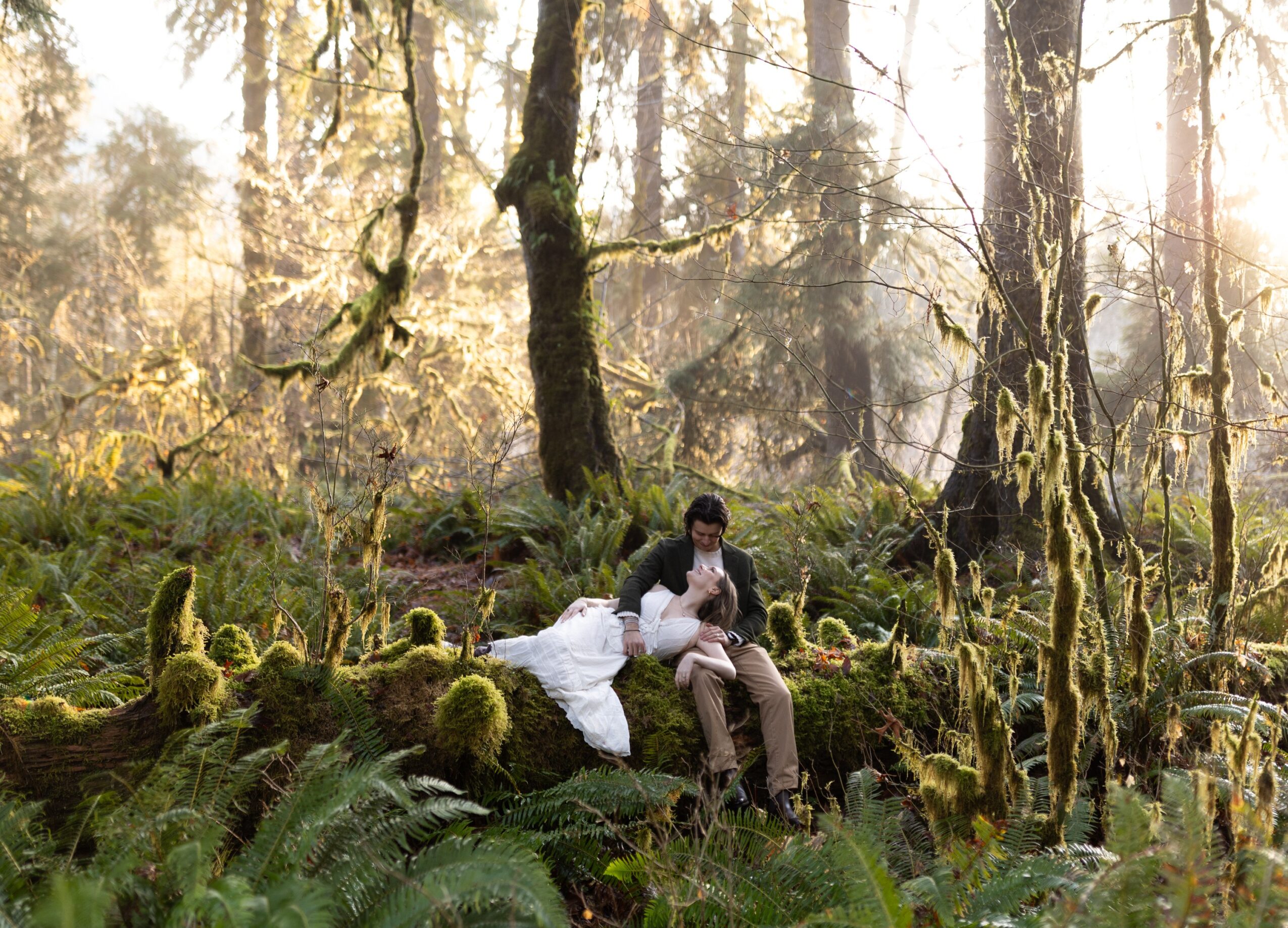 Two people sitting on a moss-covered log in a sunlit forest with tall trees and lush greenery.
