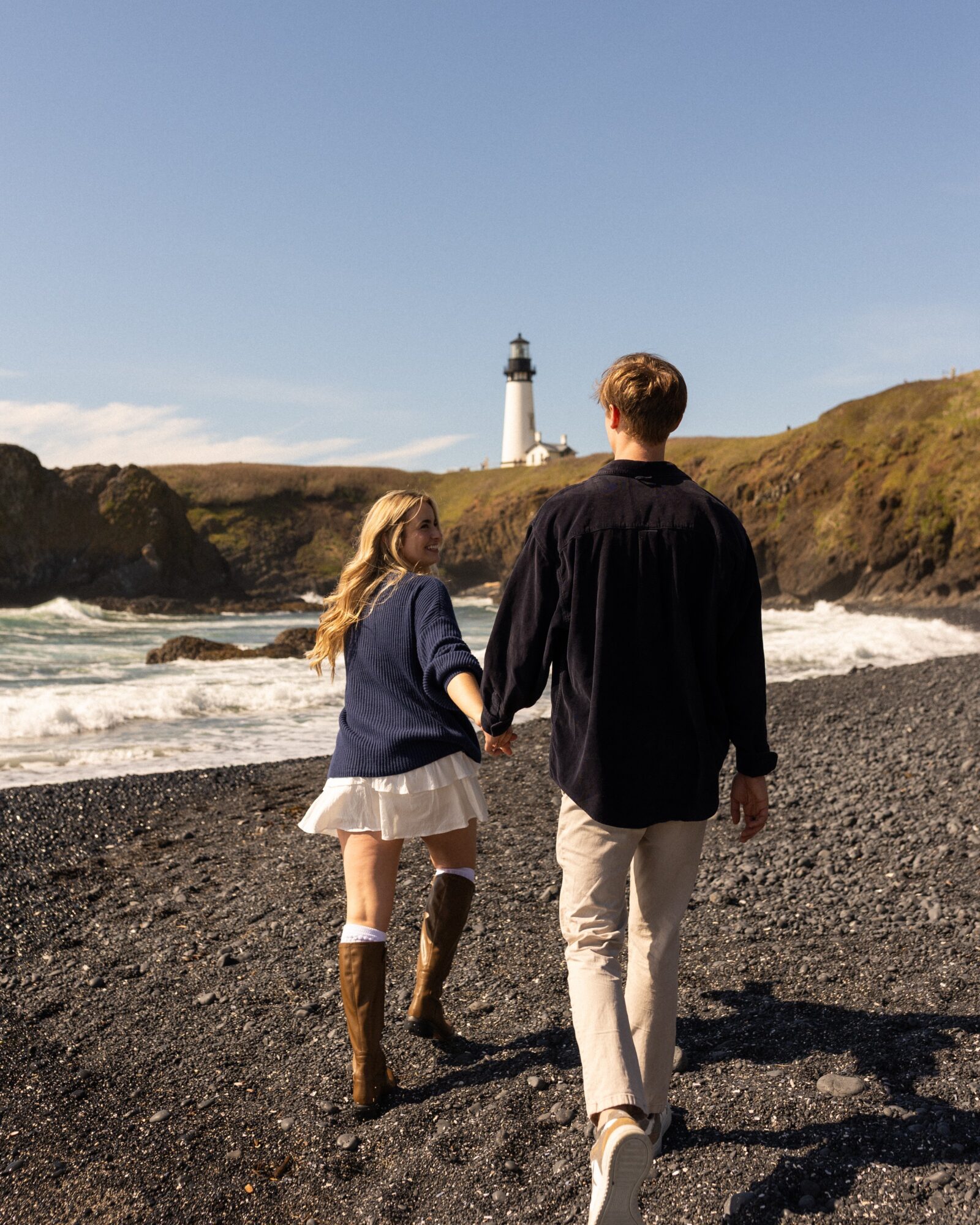 A couple holding hands walking on a rocky beach with a lighthouse in the background under a clear blue sky.