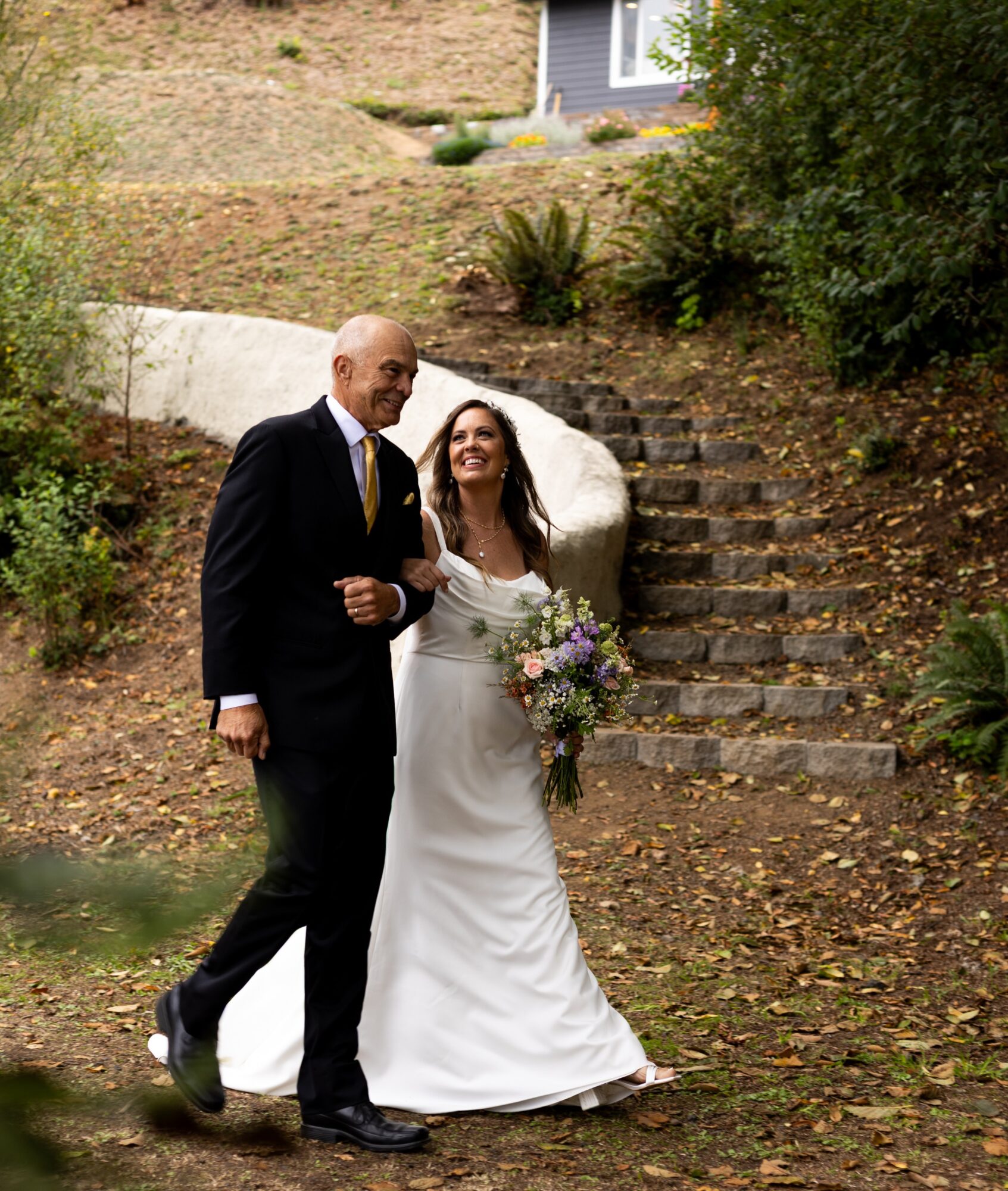 A bride and groom walking outdoors on a staircase, smiling, with trees and a house in the background.