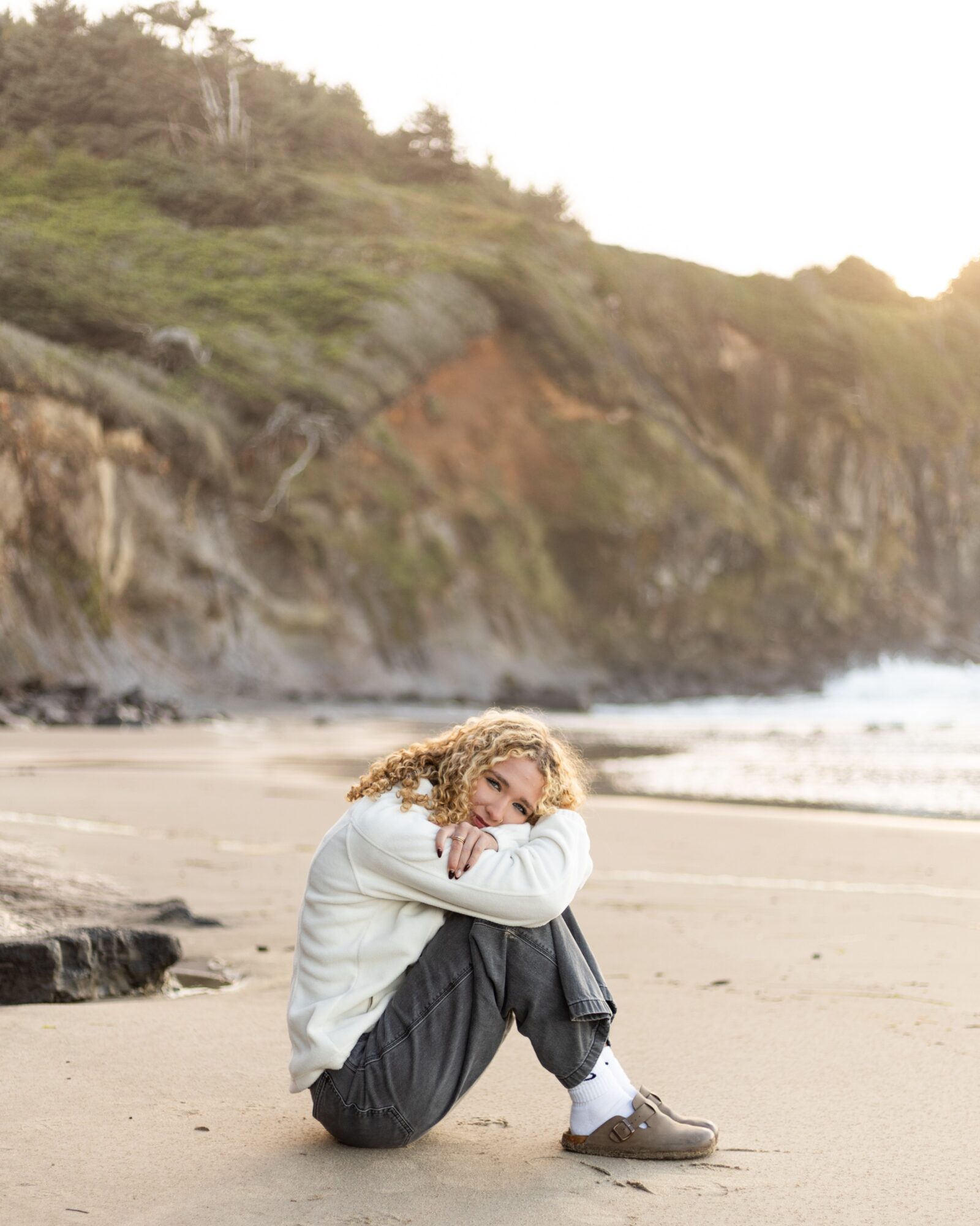 Person sitting on a beach with arms crossed, looking down, with cliffs and ocean in background
