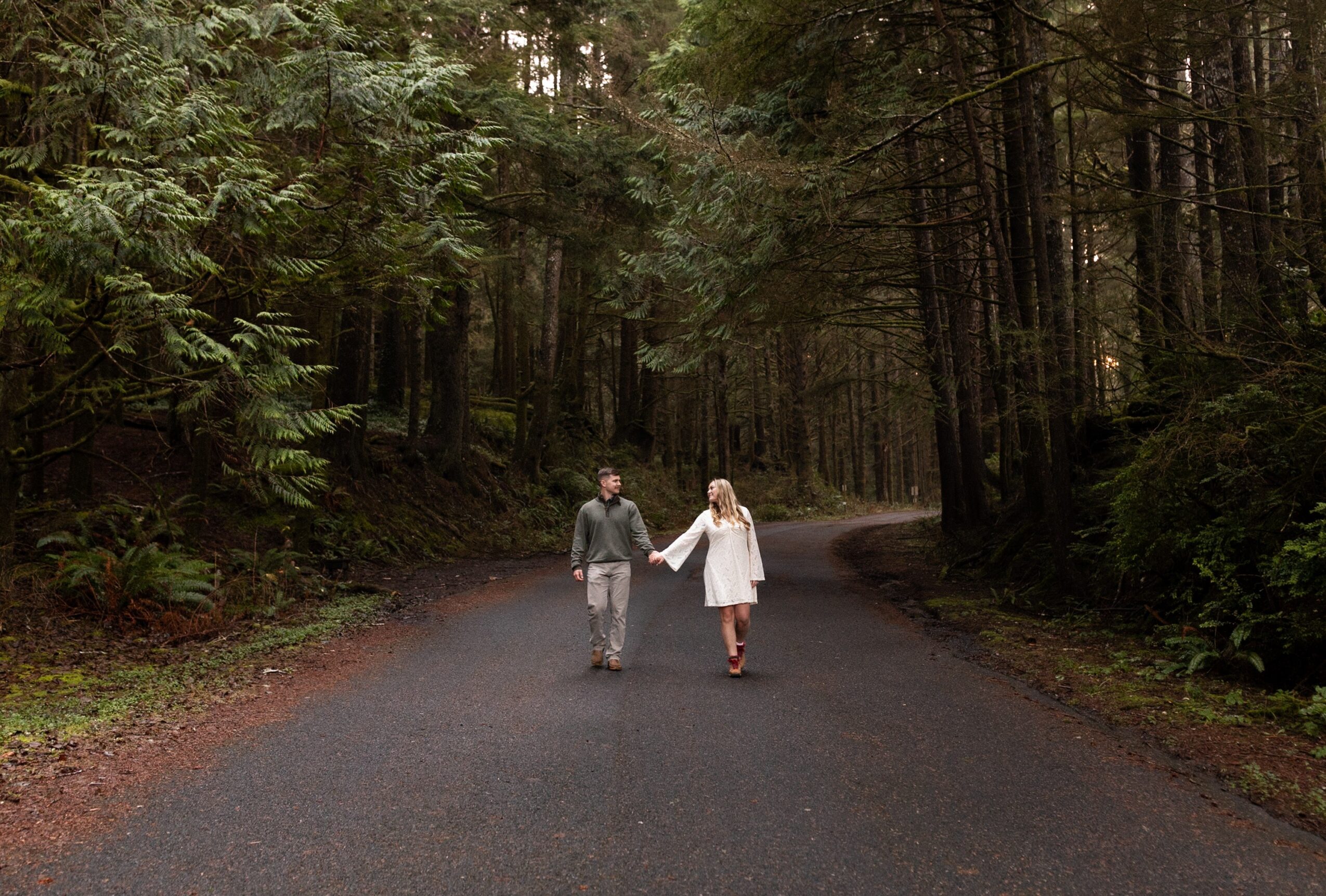A couple walks hand in hand on a forest path surrounded by tall trees, with dense greenery on both sides.