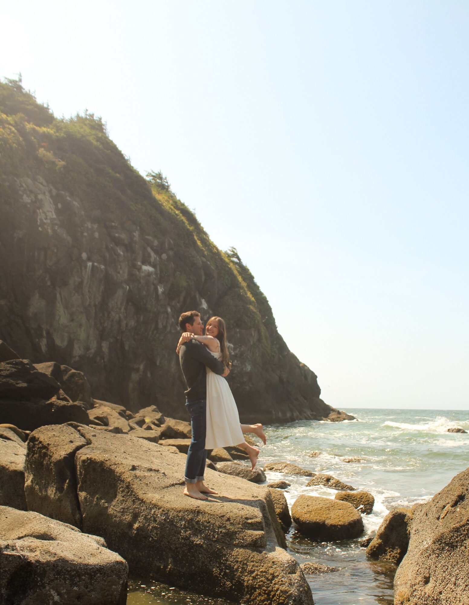 A couple stands on rocks near the ocean, embracing with a cliff in the background and a clear sky above.