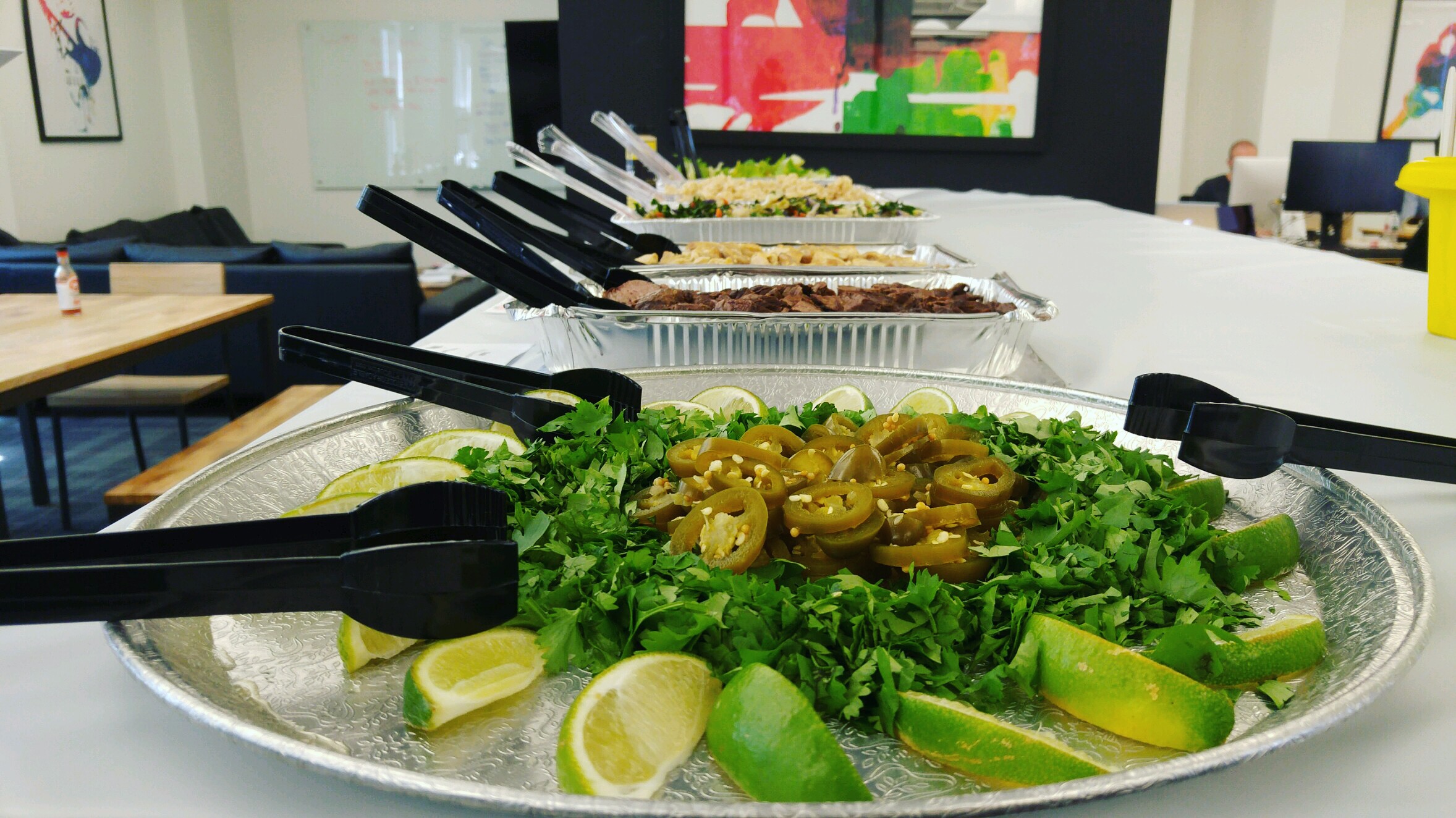 Tray of sliced green olives, lime wedges, and fresh lettuce on a table, with other trays in the background.