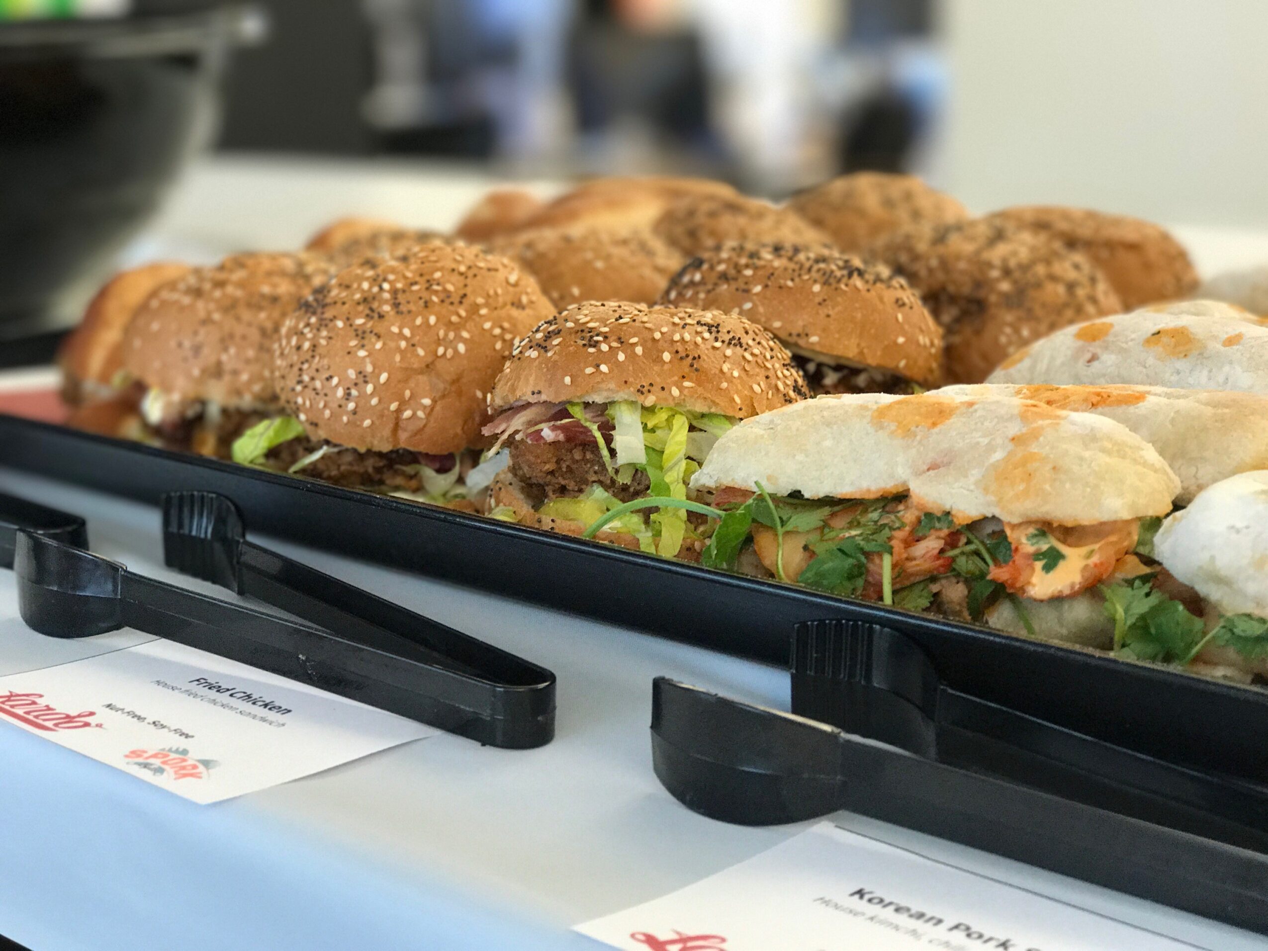 Several sandwiches with sesame seed buns and lettuce on a black tray, placed on a white table, with blurred background.
