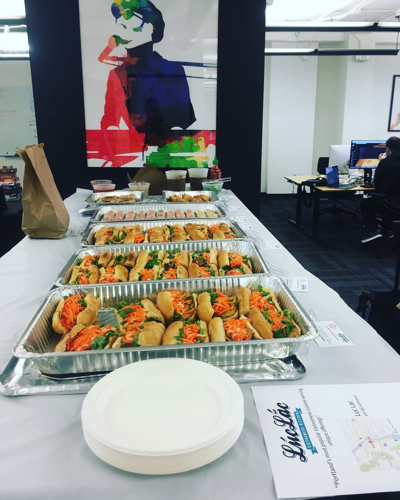 Tray of assorted sandwiches and snacks on a table with a paper plate and a menu, in a modern office setting.