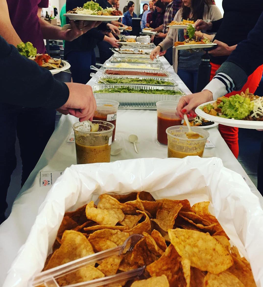 People gather around a table with food, serving salads and drinks, with a basket of chips in the foreground.