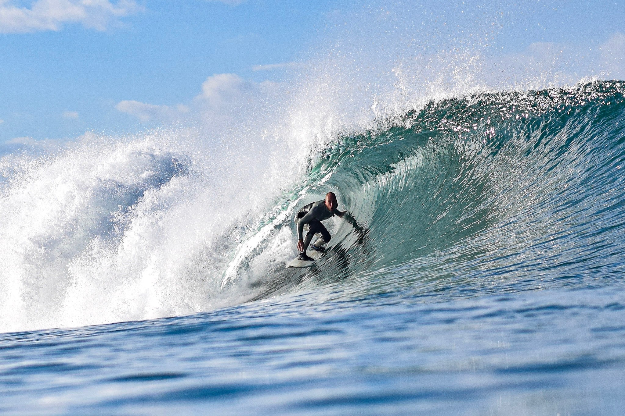 Surfer riding inside a large wave with a blue sky background, water splashing around, and a person in a wetsuit on a surfboard.