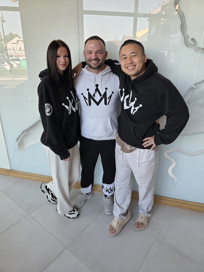 Three people standing indoors, smiling, with large windows and a mirror in the background.