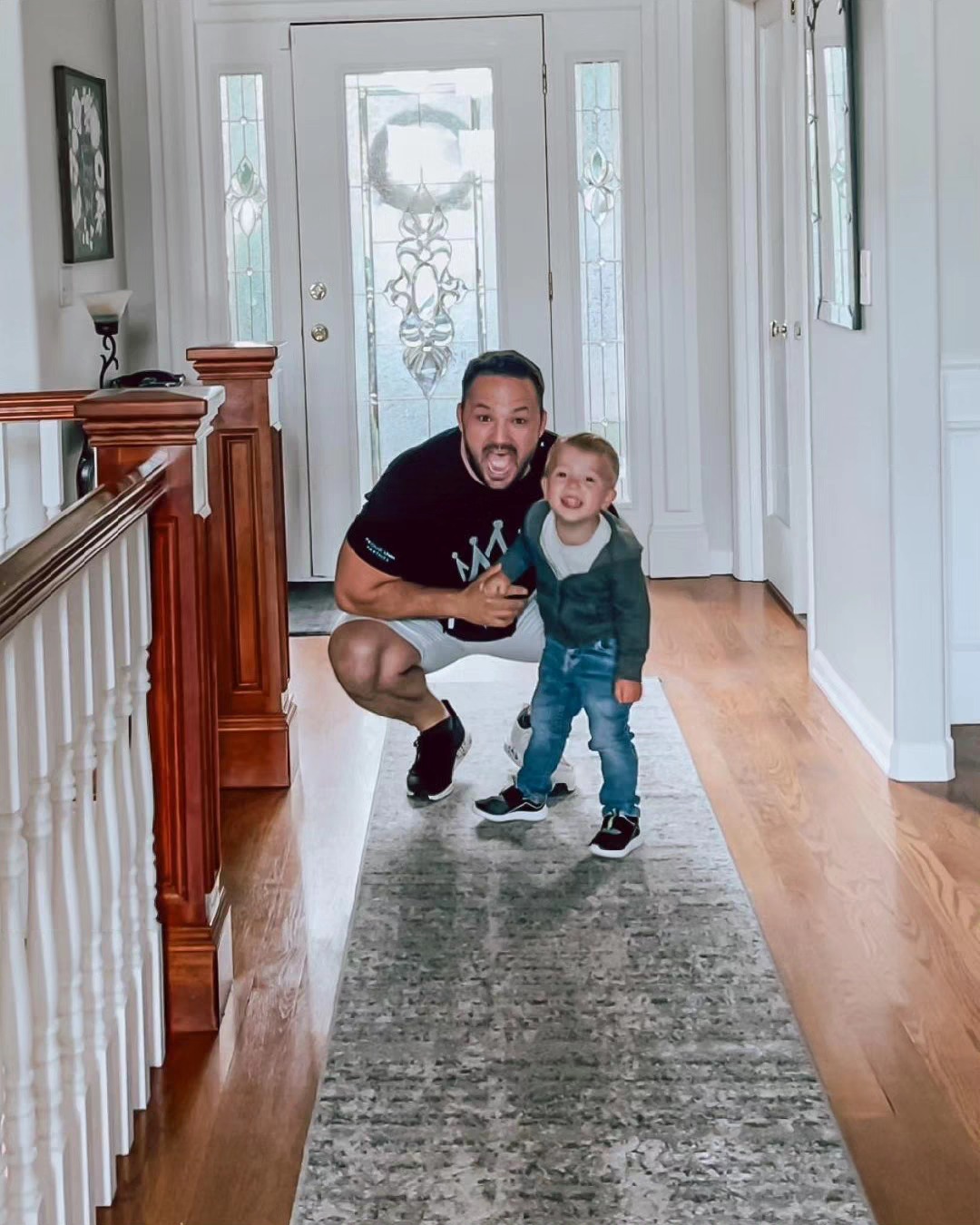 Man and young child smiling and playing in hallway with wooden floor and glass door, natural light, framed pictures on wall