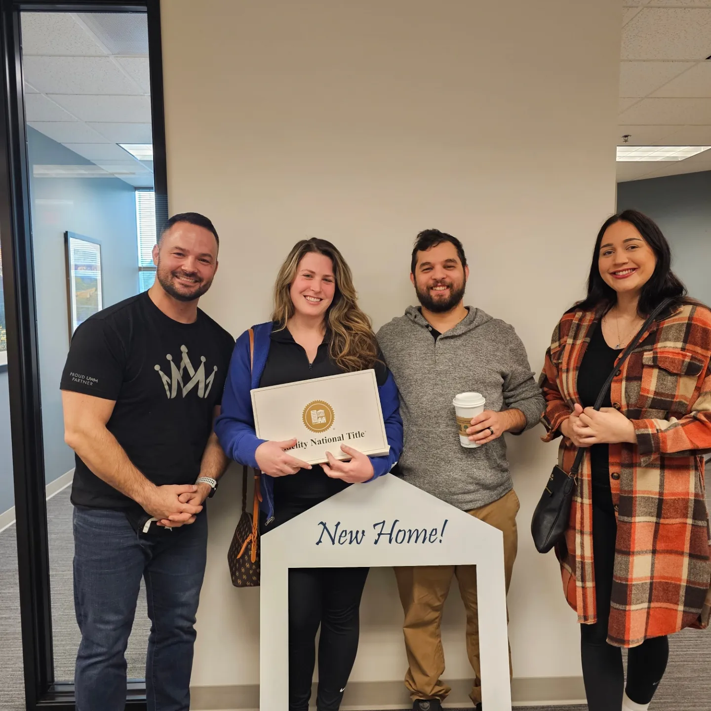 Four people standing indoors, smiling, with a sign that says