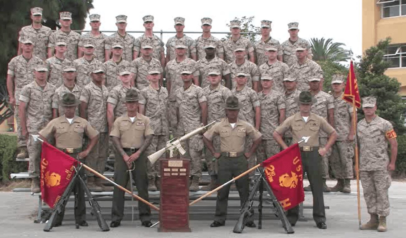 Group of soldiers in uniform standing outdoors, with flags and a monument, in a formal formation.