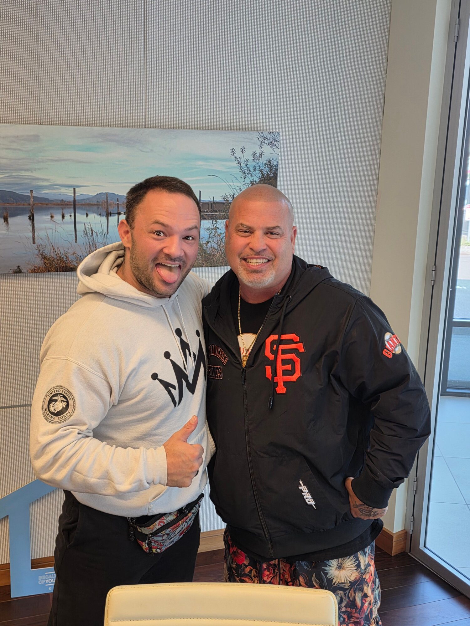 Two men standing indoors, one in a black jacket and the other in a white hoodie, smiling at the camera.