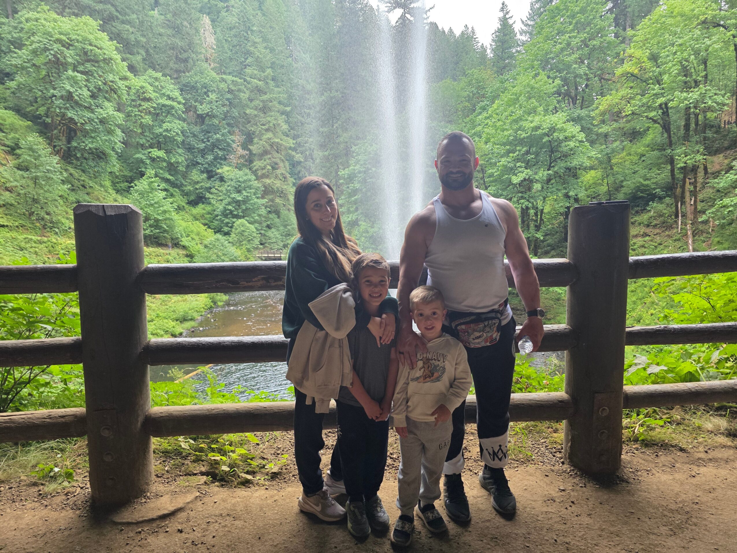 Group of four people standing in front of a waterfall with lush green trees, behind a wooden fence.