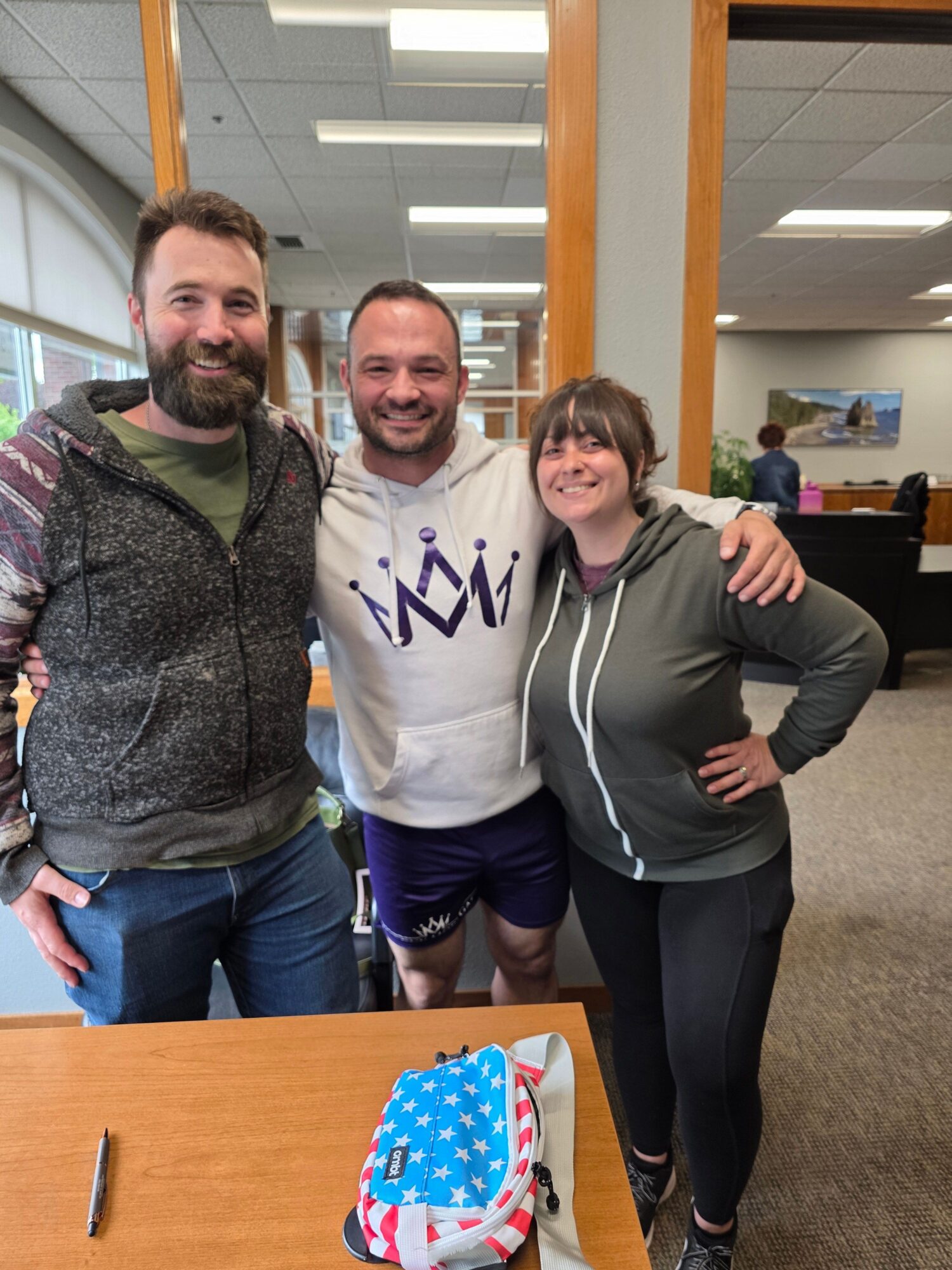 Three people standing indoors, smiling, with a table and a backpack nearby.
