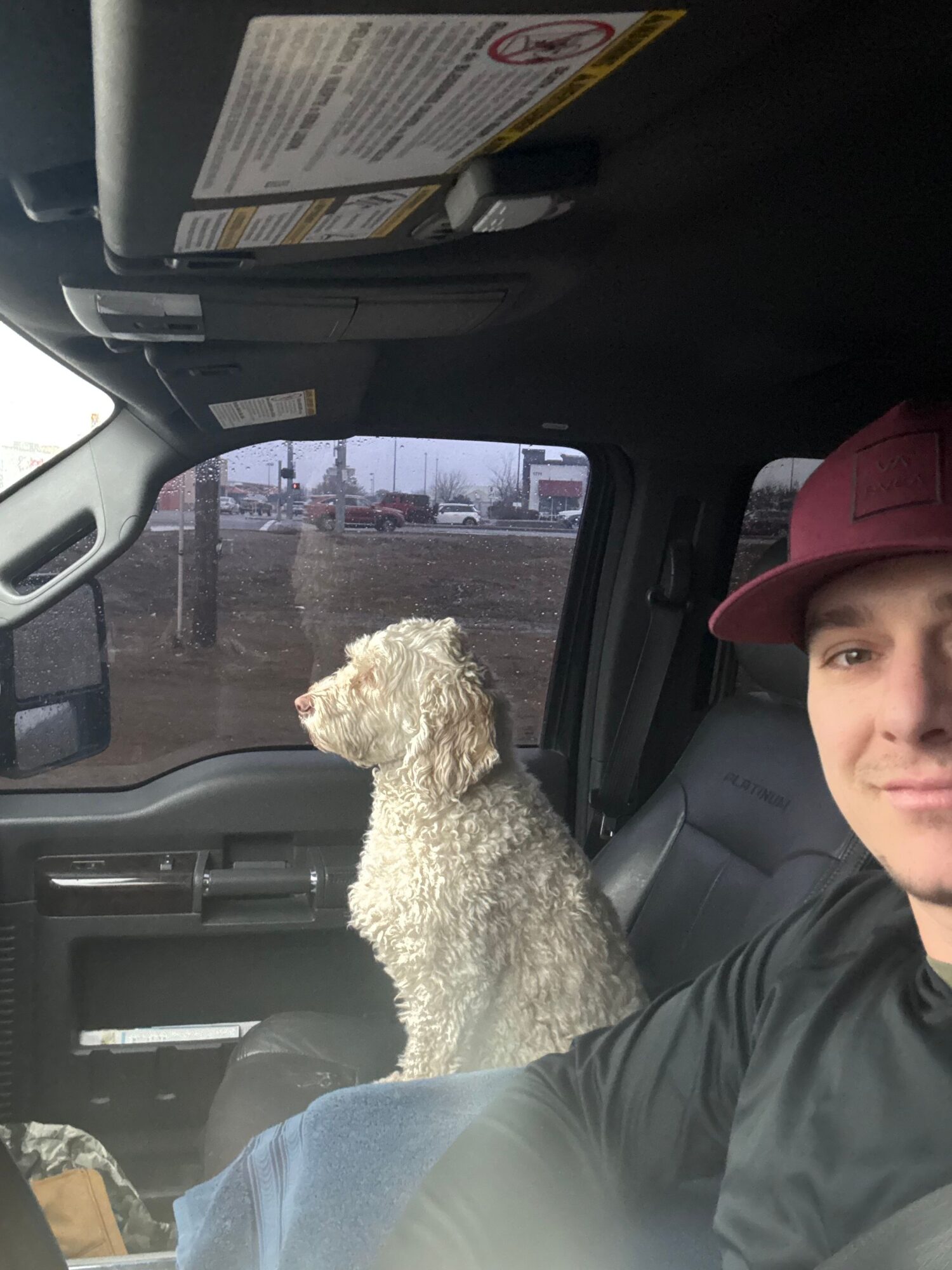 Person wearing a maroon cap sitting in a vehicle with a curly-haired dog beside them, outside view through window.