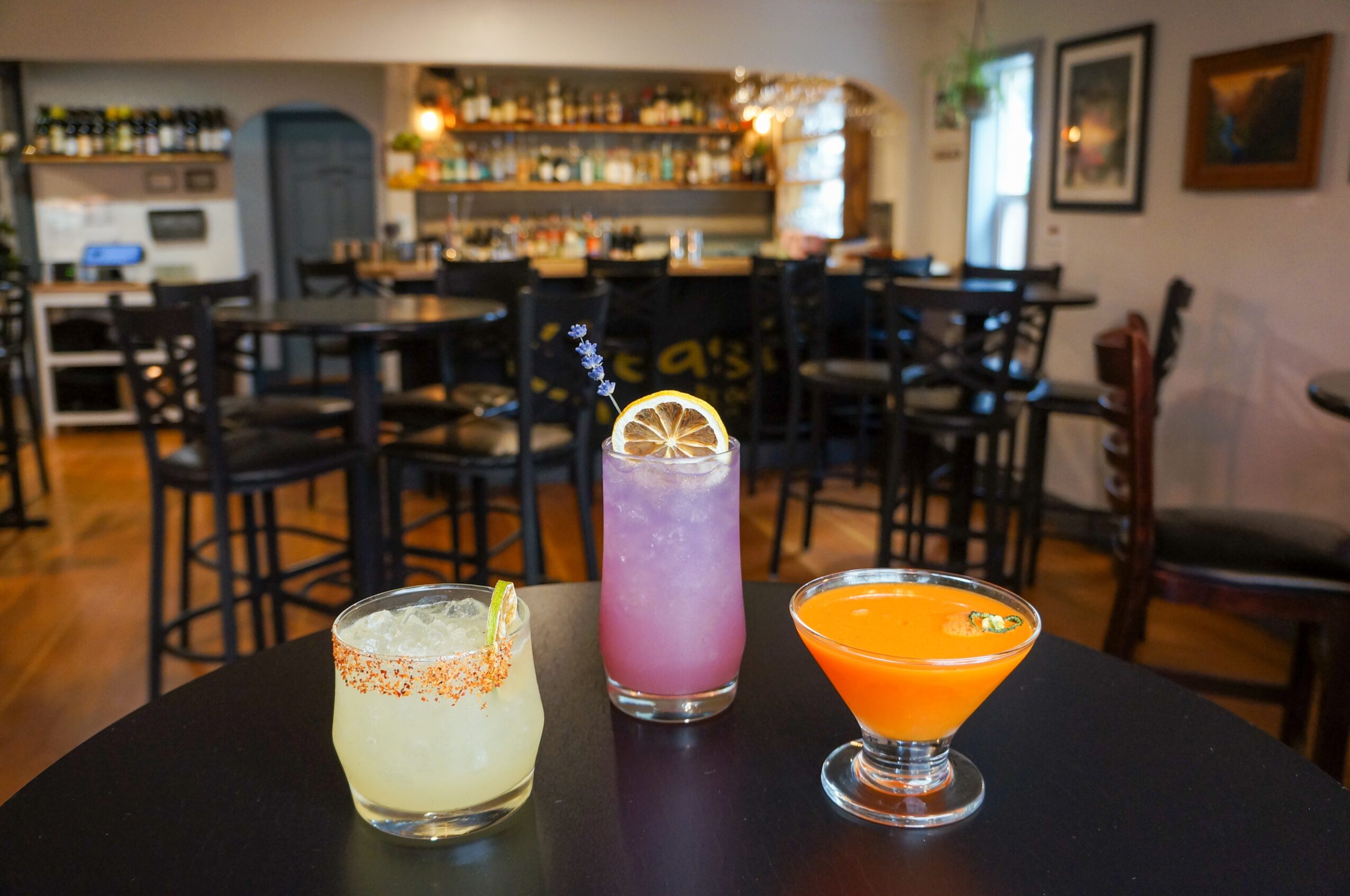 Three colorful cocktails on a table in a bar with chairs and artwork in the background.