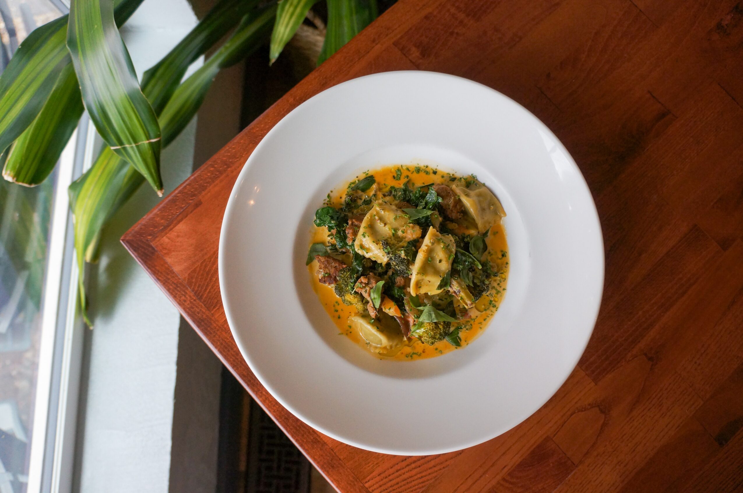 Bowl of soup with vegetables and herbs on a wooden table, viewed from above.