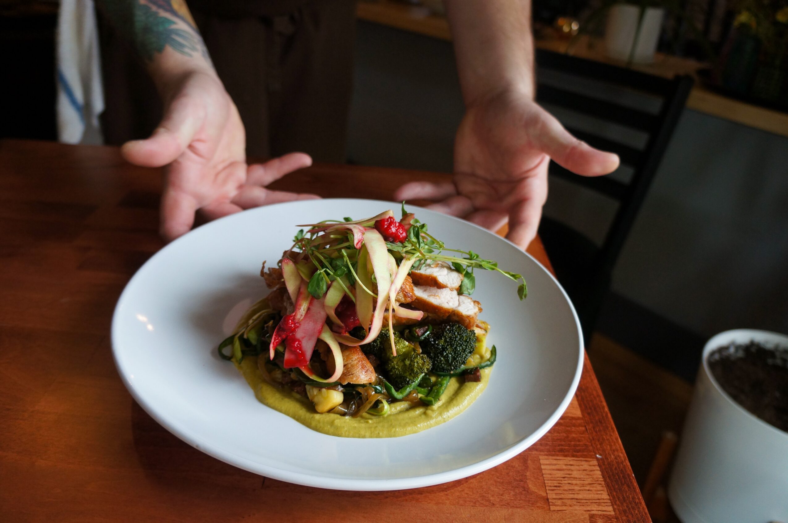 Person presenting a plate of pasta with vegetables and herbs on a wooden table, with a drink nearby.