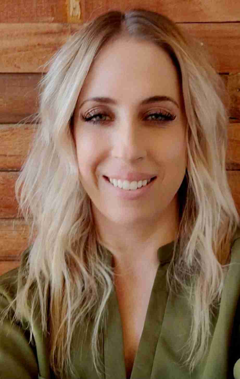 Smiling woman with wavy blonde hair and light makeup, wearing a green top, in front of wooden background.