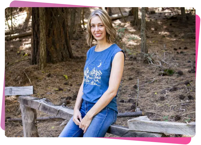 Young woman with blonde hair smiling outdoors, wearing a blue sleeveless top and jeans, sitting on a wooden fence in a forest.