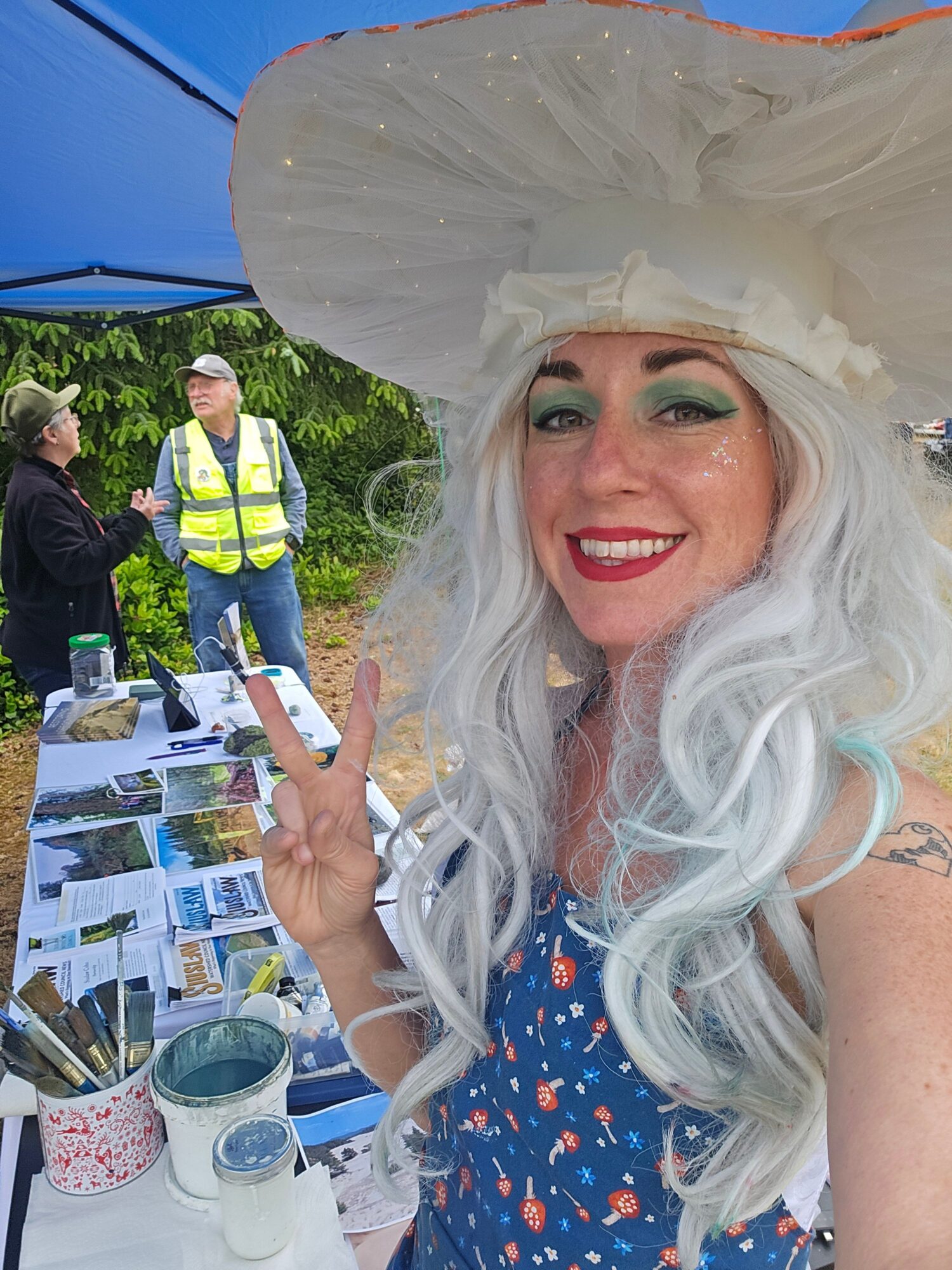 Woman with long white hair and makeup, wearing a large hat, smiling outdoors near a table with papers and bottles.