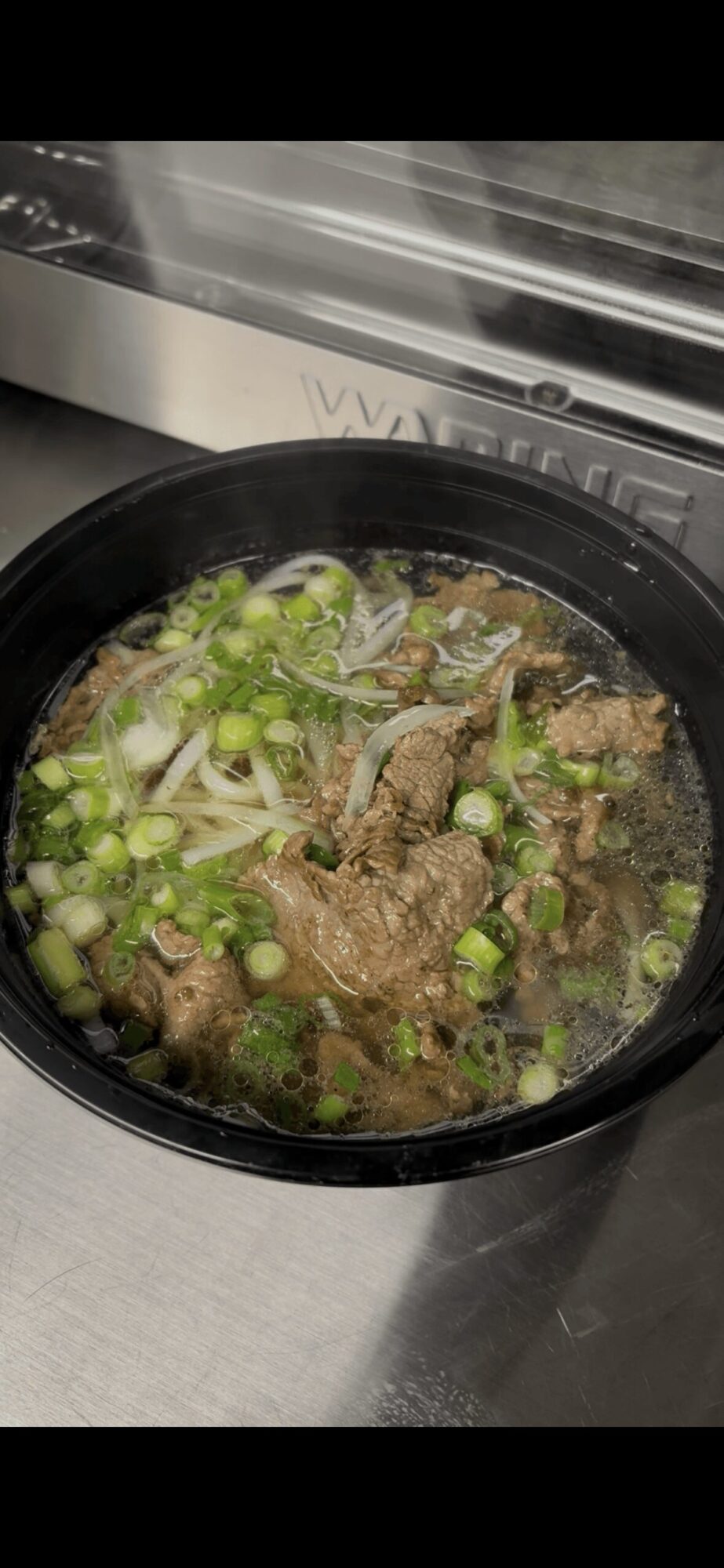 Soup with meat and chopped green onions in a black bowl, placed on a metal surface near a window.