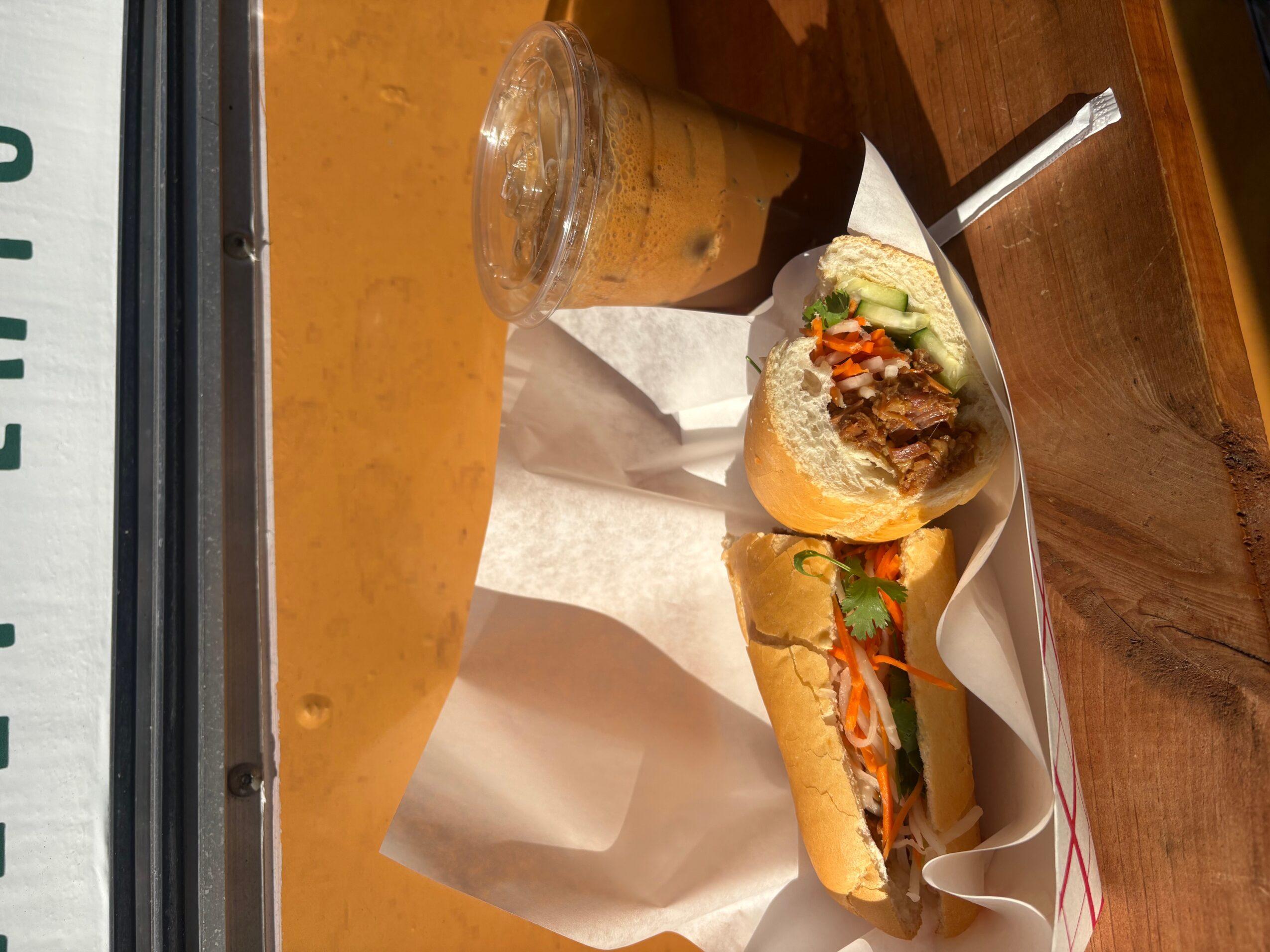 Sandwich with vegetables and meat, a drink, and a dessert on a wooden table.