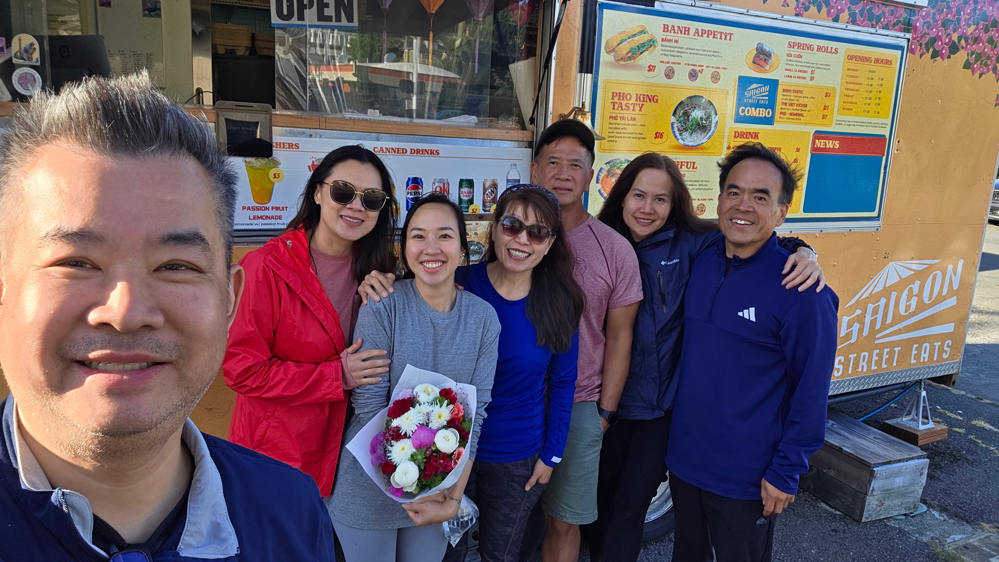 Group of six people standing outside a food truck, smiling, with one person holding a bouquet of flowers.