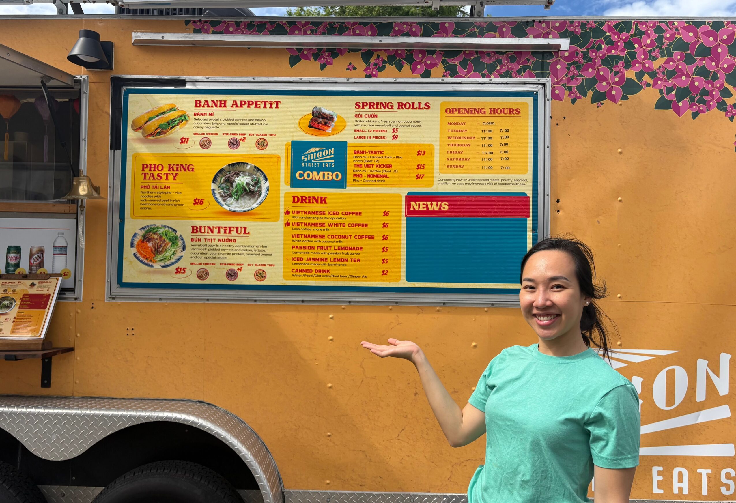 Woman smiling and pointing at a colorful food menu board outdoors, with a yellow wall and pink flowers in the background.