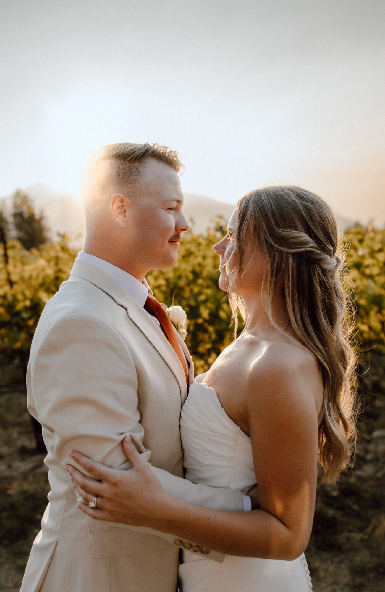A couple stands close outdoors, gazing at each other, with trees and sky in the background.