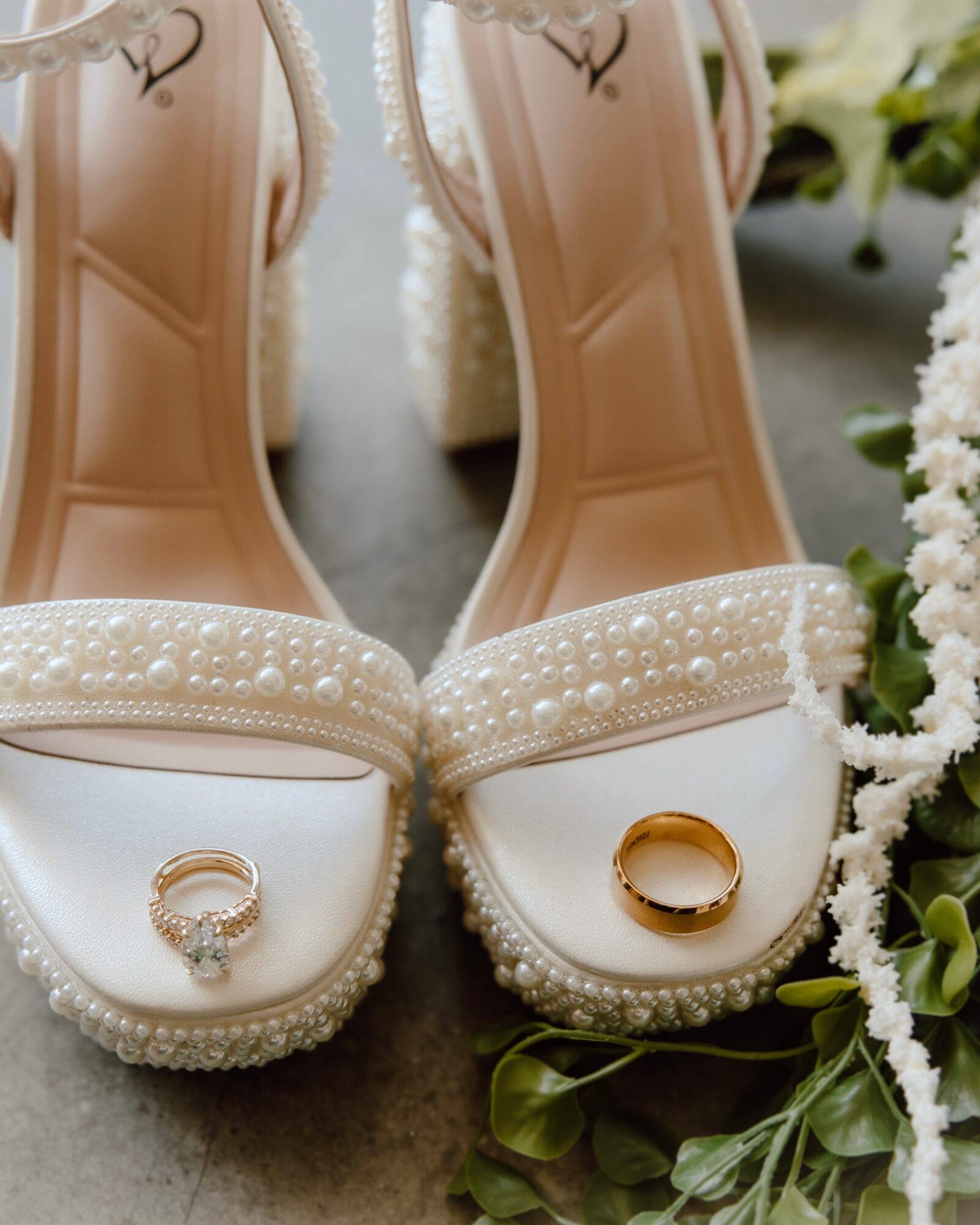 Pair of white high-heeled shoes with pearl embellishments, each holding a ring, surrounded by white flowers and greenery.