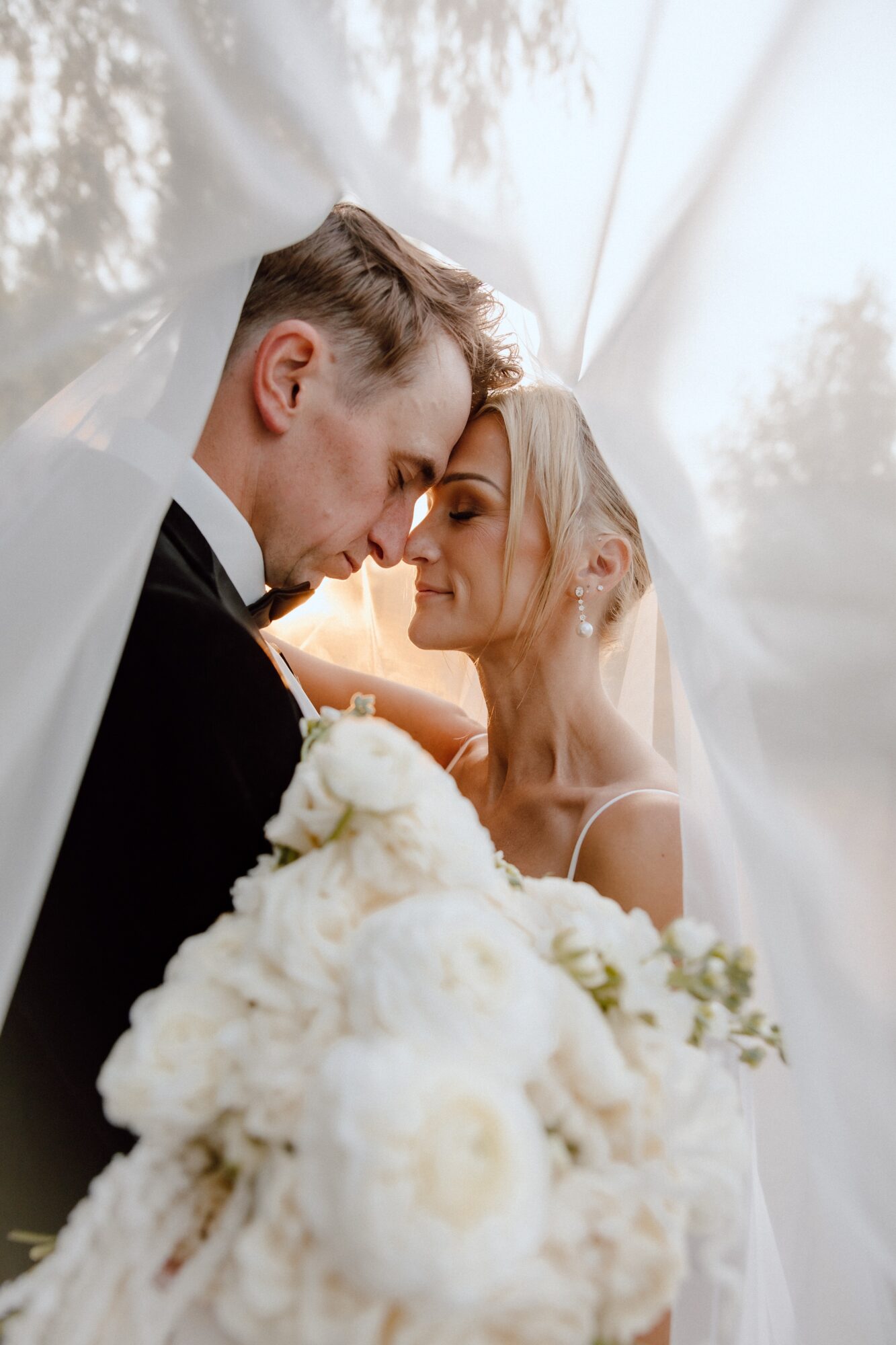 A bride and groom with foreheads touching, holding a bouquet of white flowers, under a white canopy.