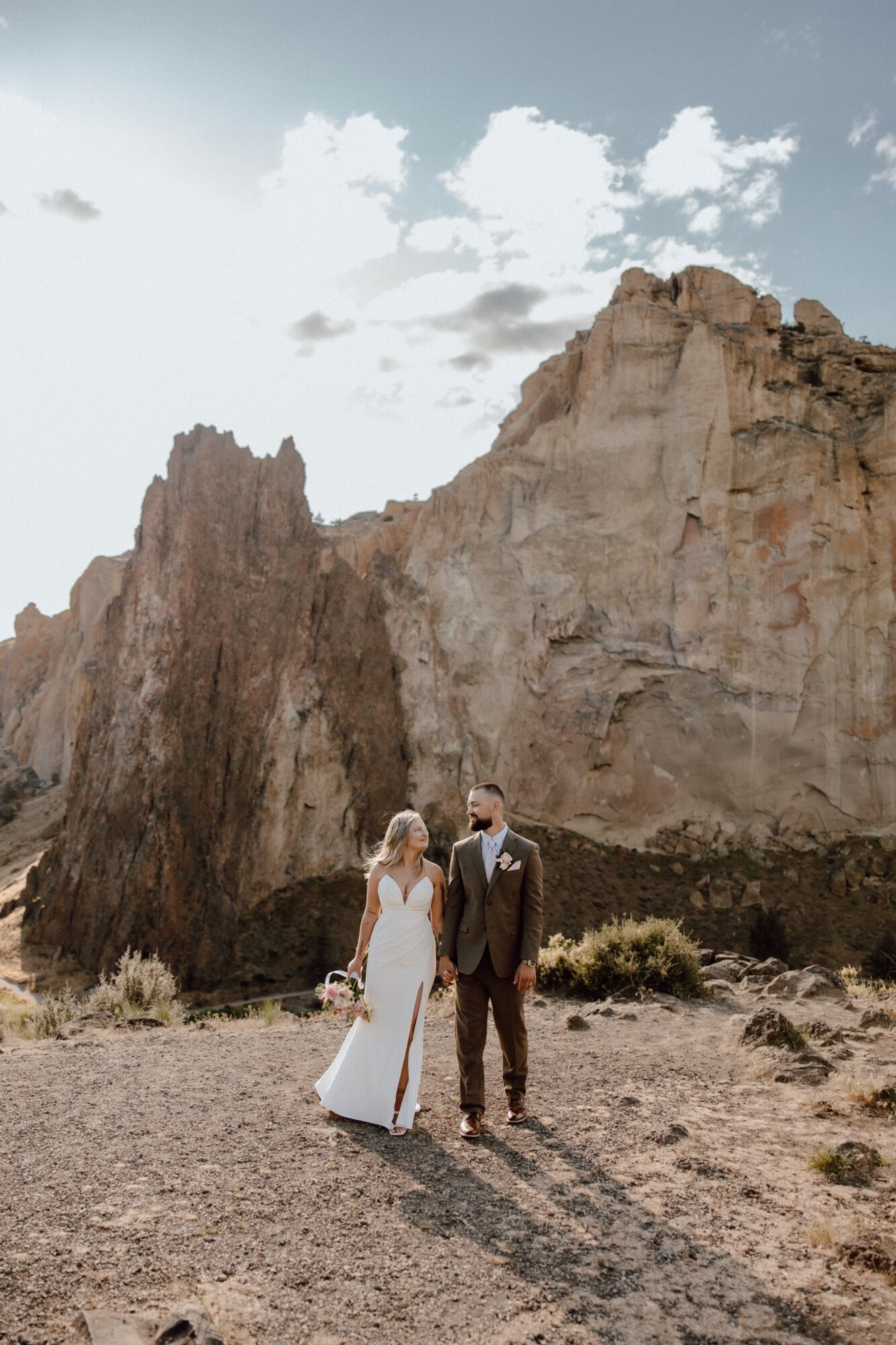 A couple in wedding attire standing outdoors with large rocky cliffs and a partly cloudy sky in the background.
