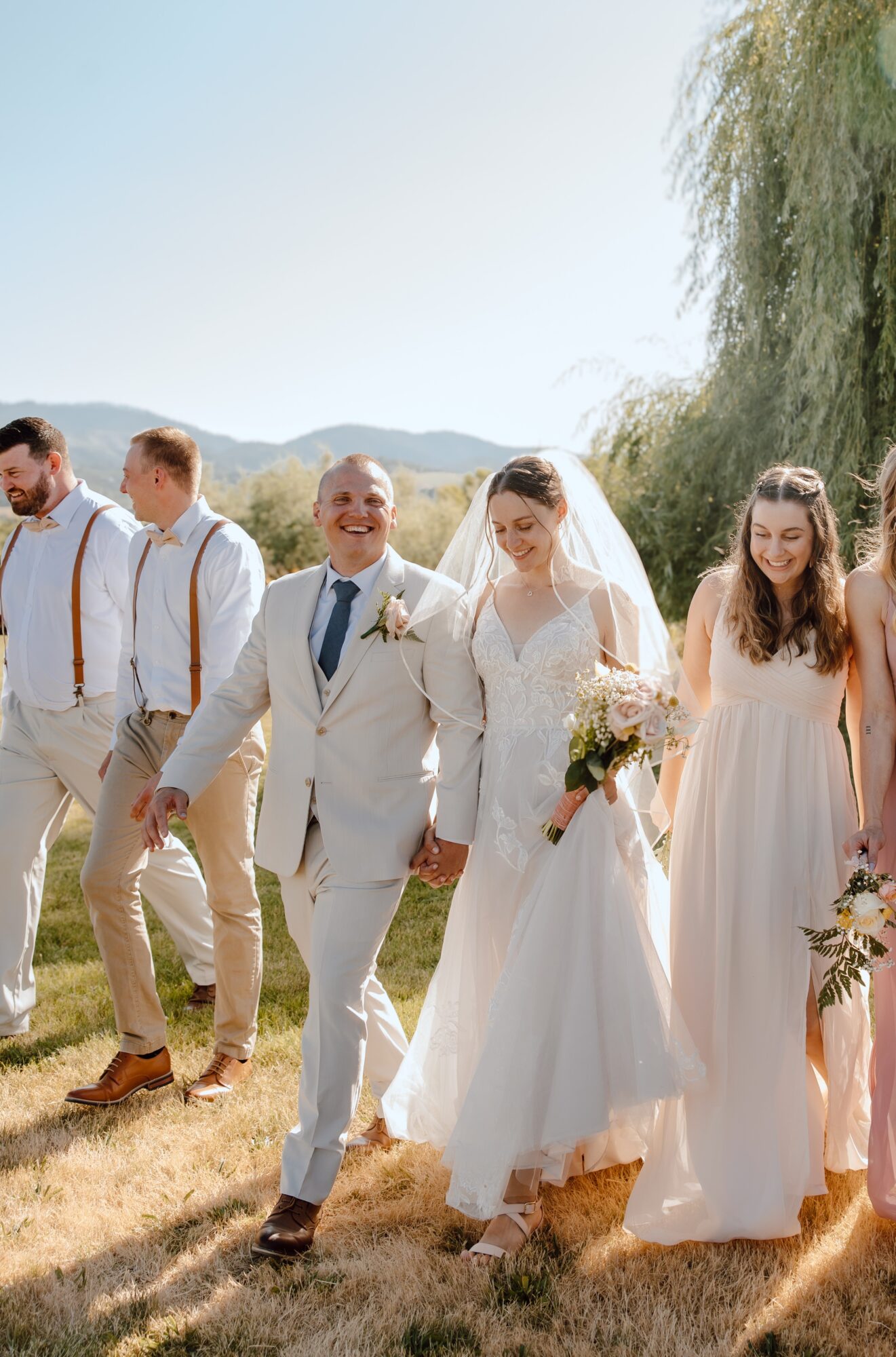 Group of people outdoors, including a bride and groom, walking on grass with trees and mountains in background.