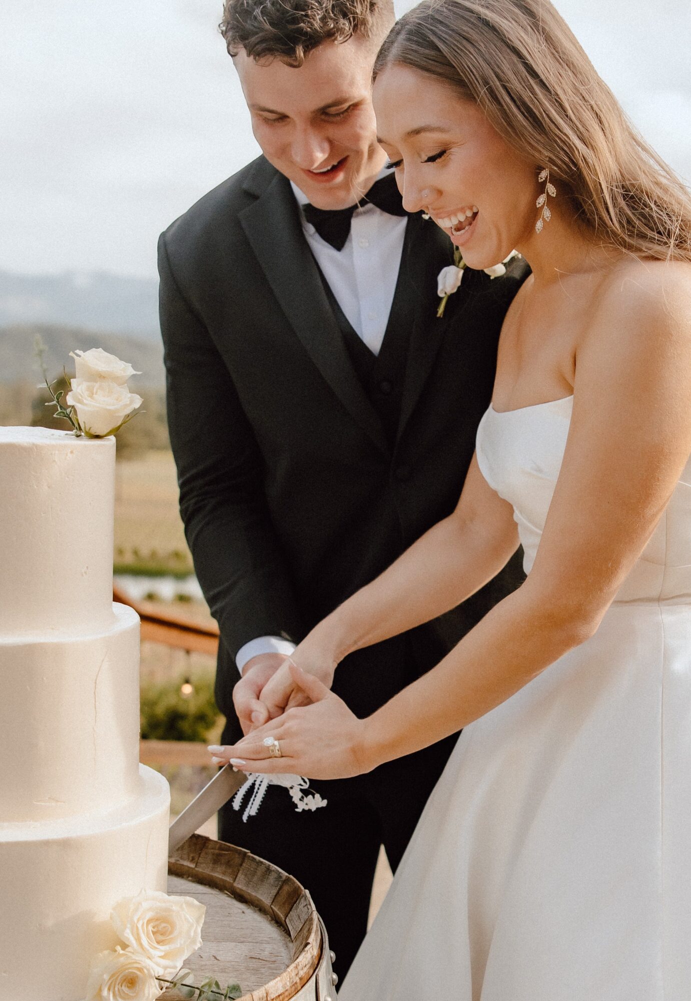 A bride and groom cut a wedding cake outdoors, smiling and holding a knife together.