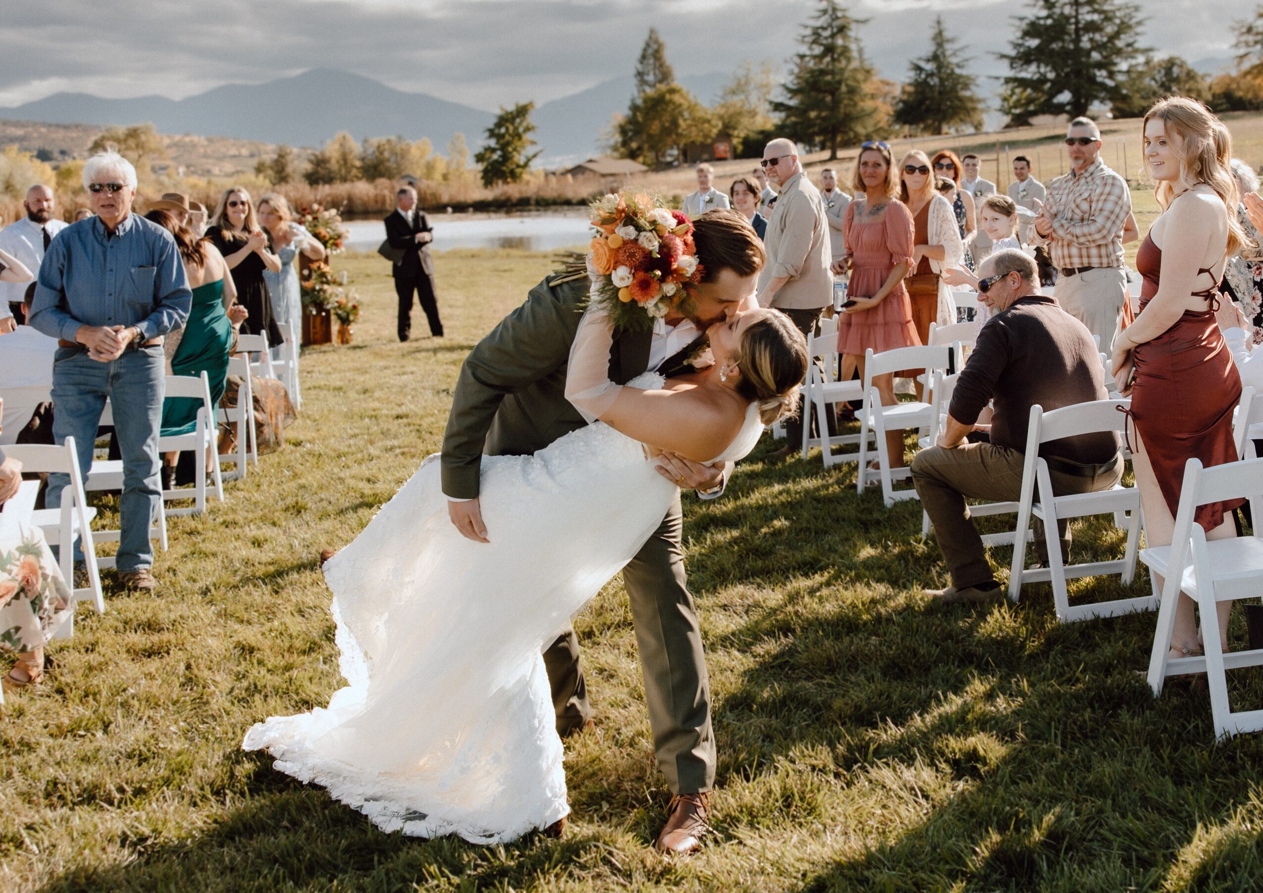 Couple dancing outdoors with guests seated and standing, scenic landscape with mountains in background, sunny weather.